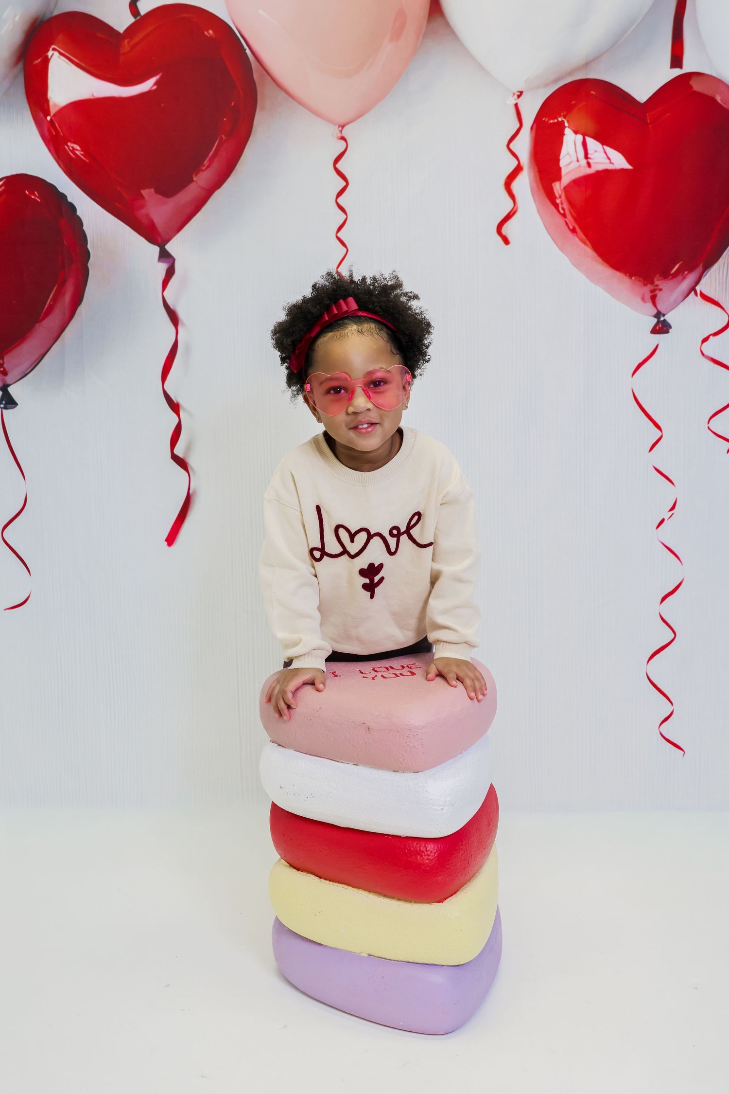 Young girl in love sweatshirt poses with stacked heart boxes and red heart balloons for Valentine's Day photo.