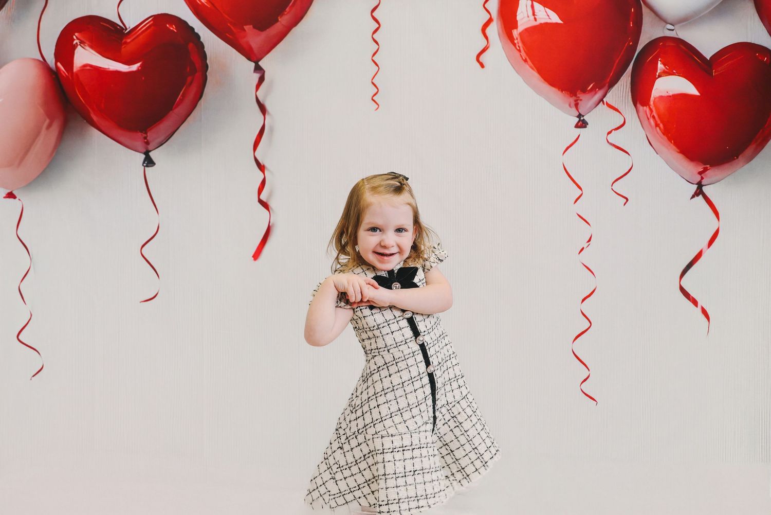 Young girl in plaid dress smiling and dancing among red and pink heart-shaped Valentine's Day balloons.