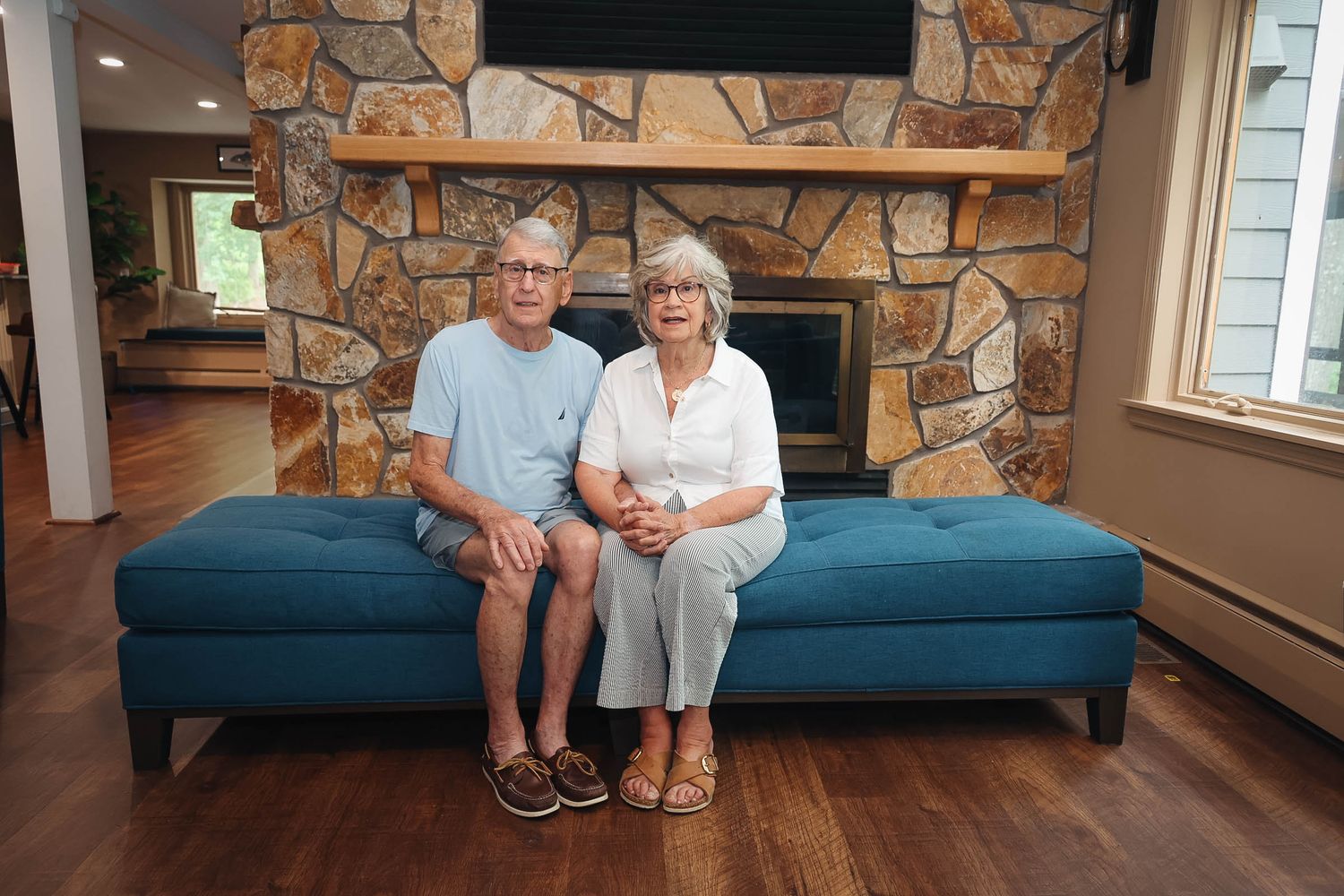 Elderly couple sitting together on a teal ottoman in front of a stone fireplace in a cozy living room.