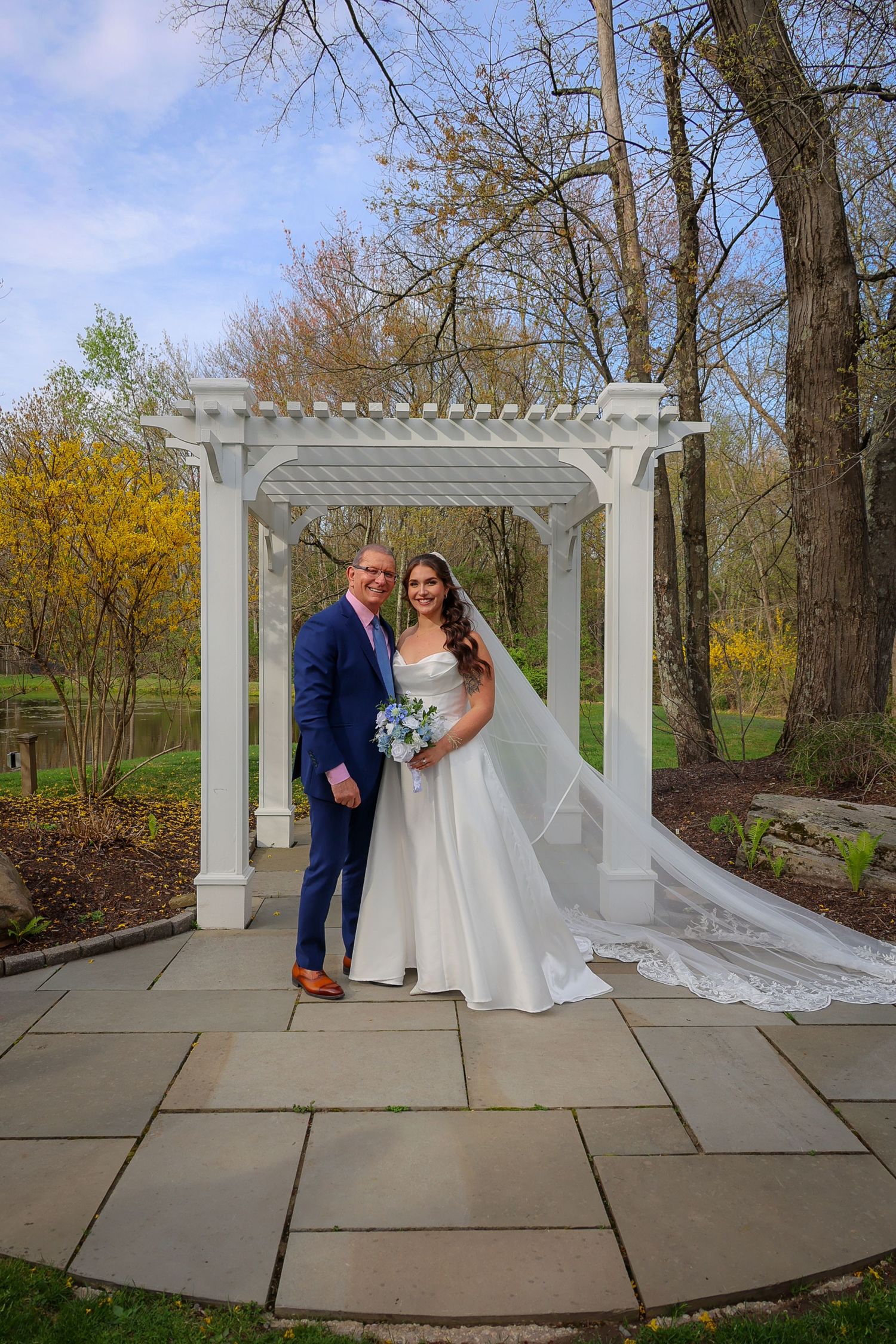 Bride and groom pose under white pergola in garden setting, bride in flowing gown with veil, groom in navy suit.