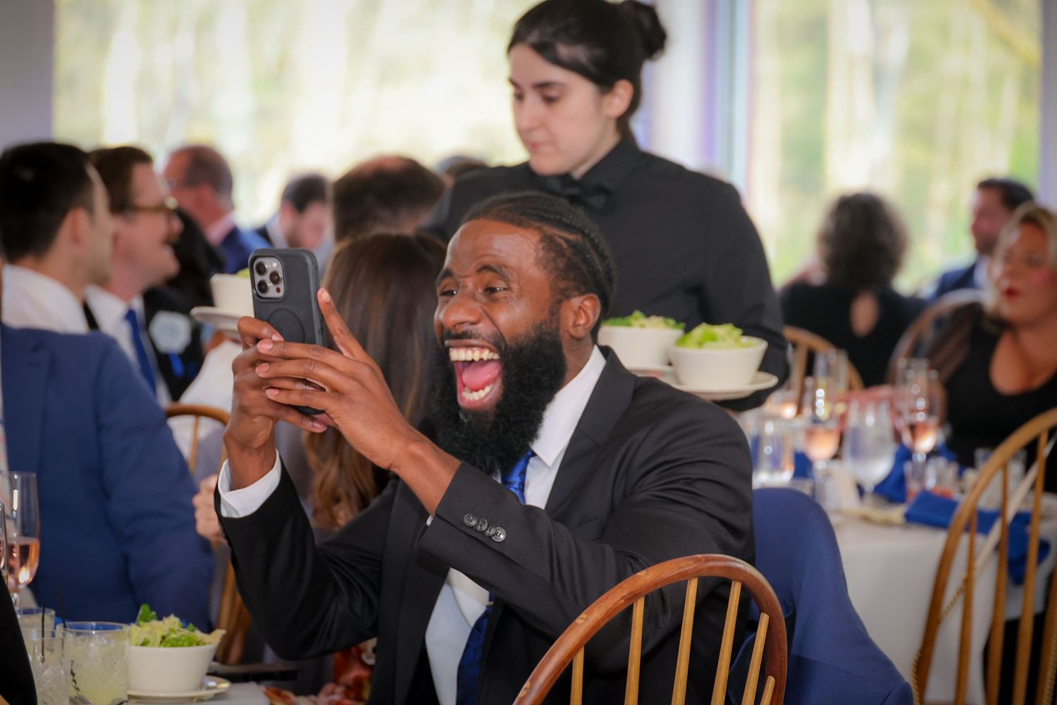 Man laughing and taking selfie at formal dinner event with guests and waiter in background.