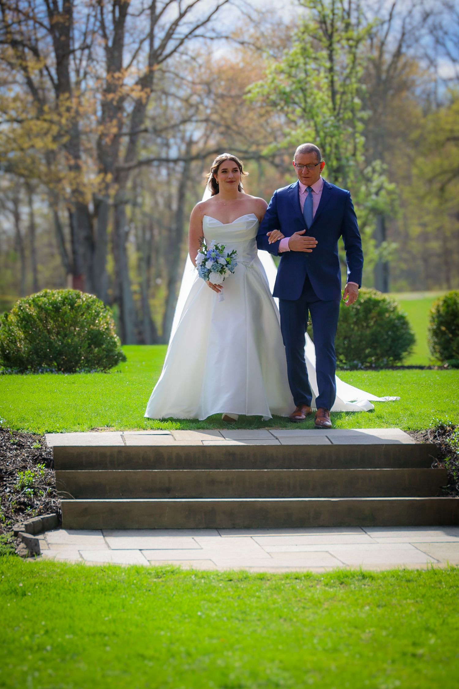Bride in white gown holding bouquet walks arm-in-arm with man in blue suit down garden steps.