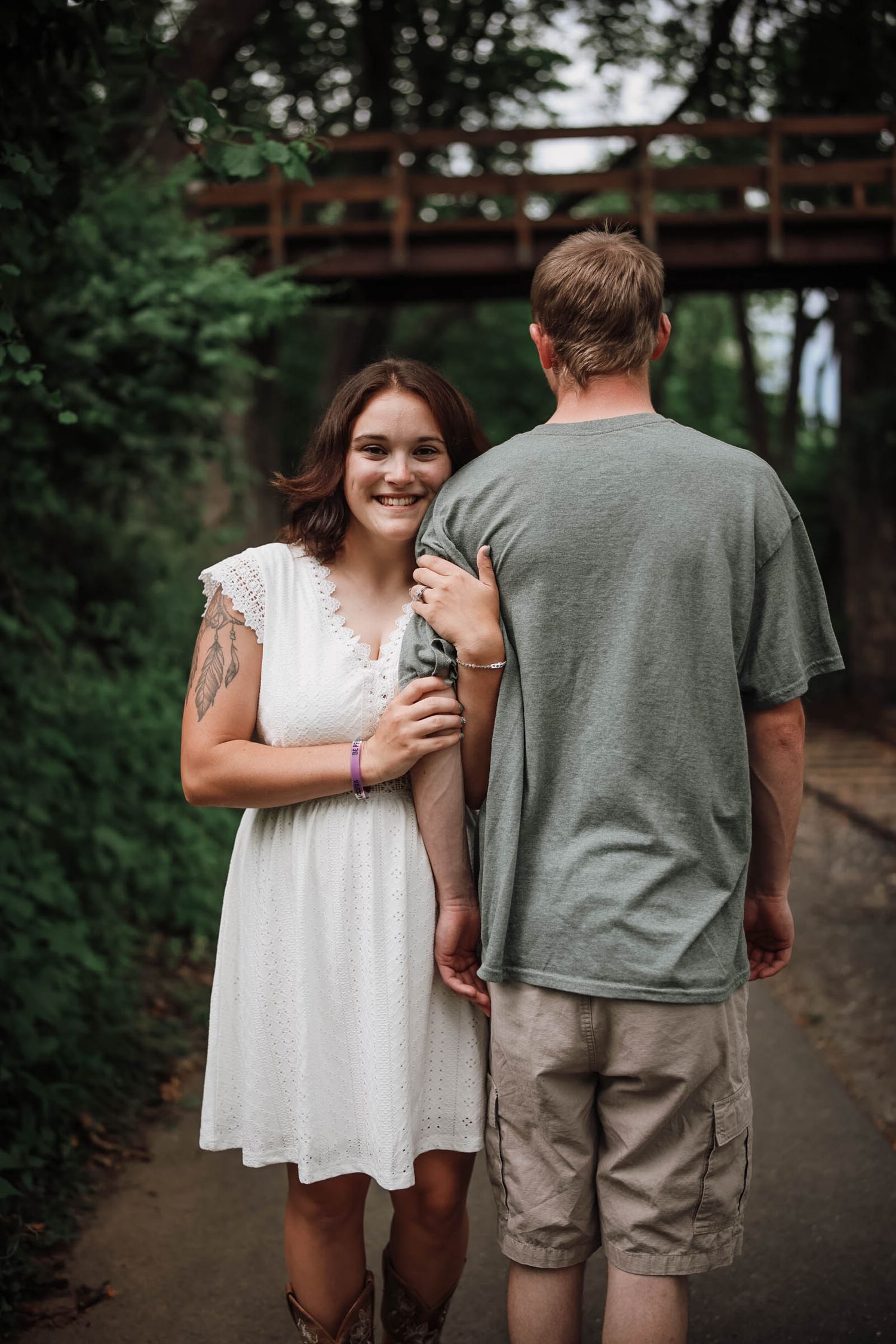 A smiling woman in a white dress holds onto a man's arm on a tree-lined path near a wooden bridge.