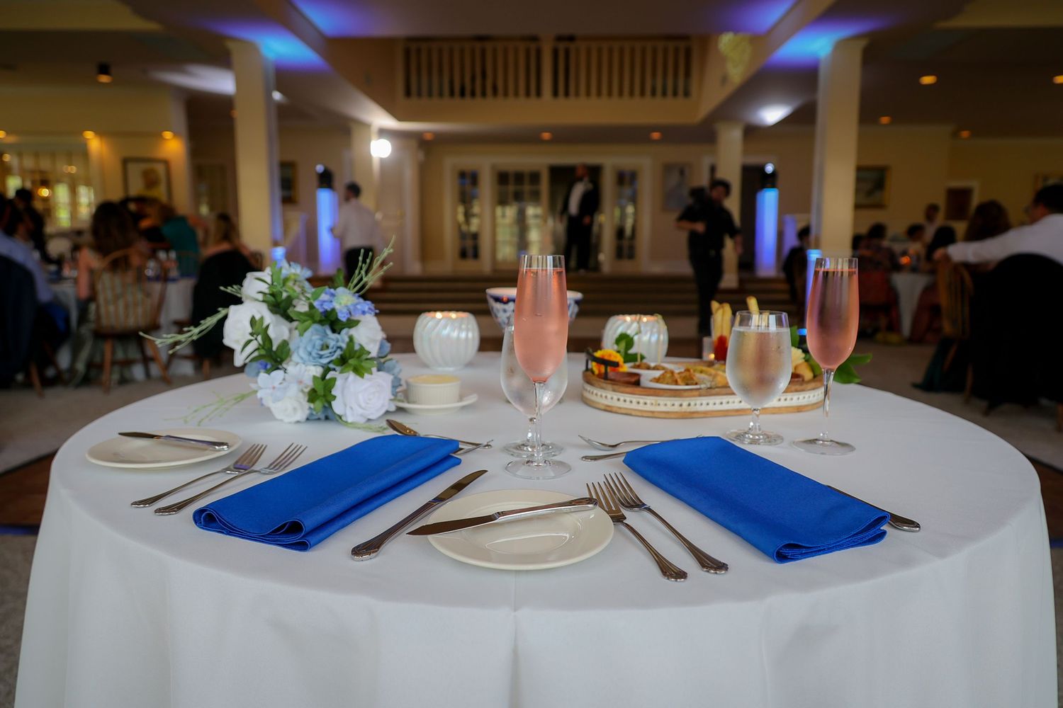 Elegant wedding reception table with blue napkins, white floral centerpiece, pink drinks, and place settings in a banquet hall.