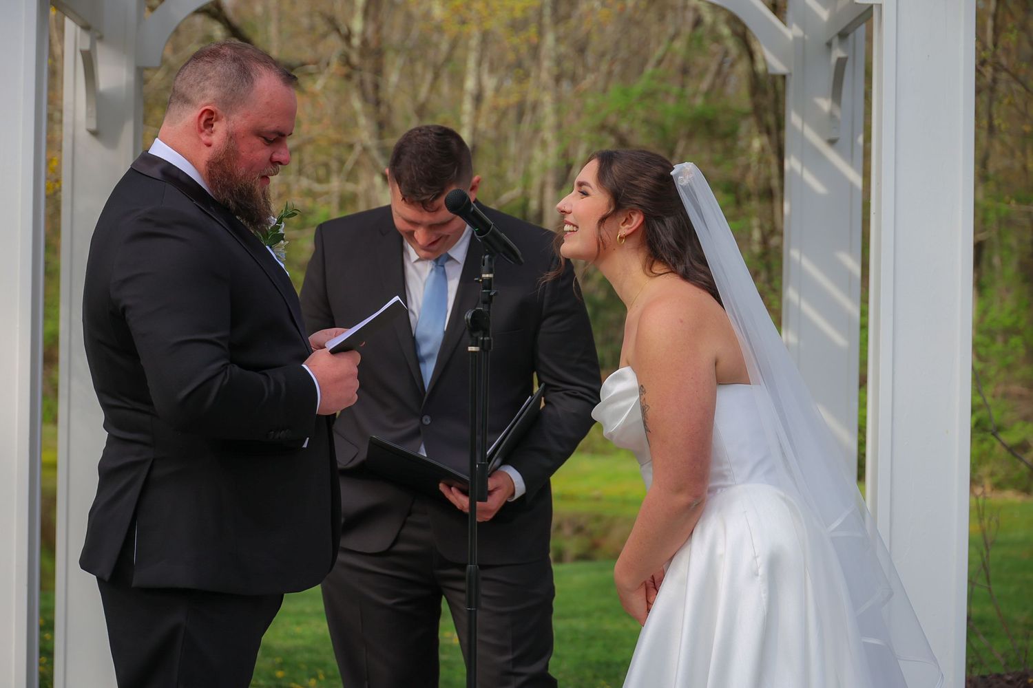 Bride and groom exchange vows with officiant at outdoor wedding ceremony under white arbor.