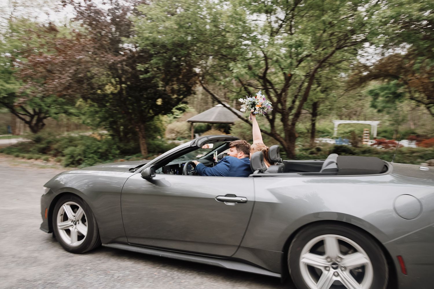 Newlyweds celebrating in a gray convertible sports car, bride holding bouquet aloft, driving through tree-lined road.