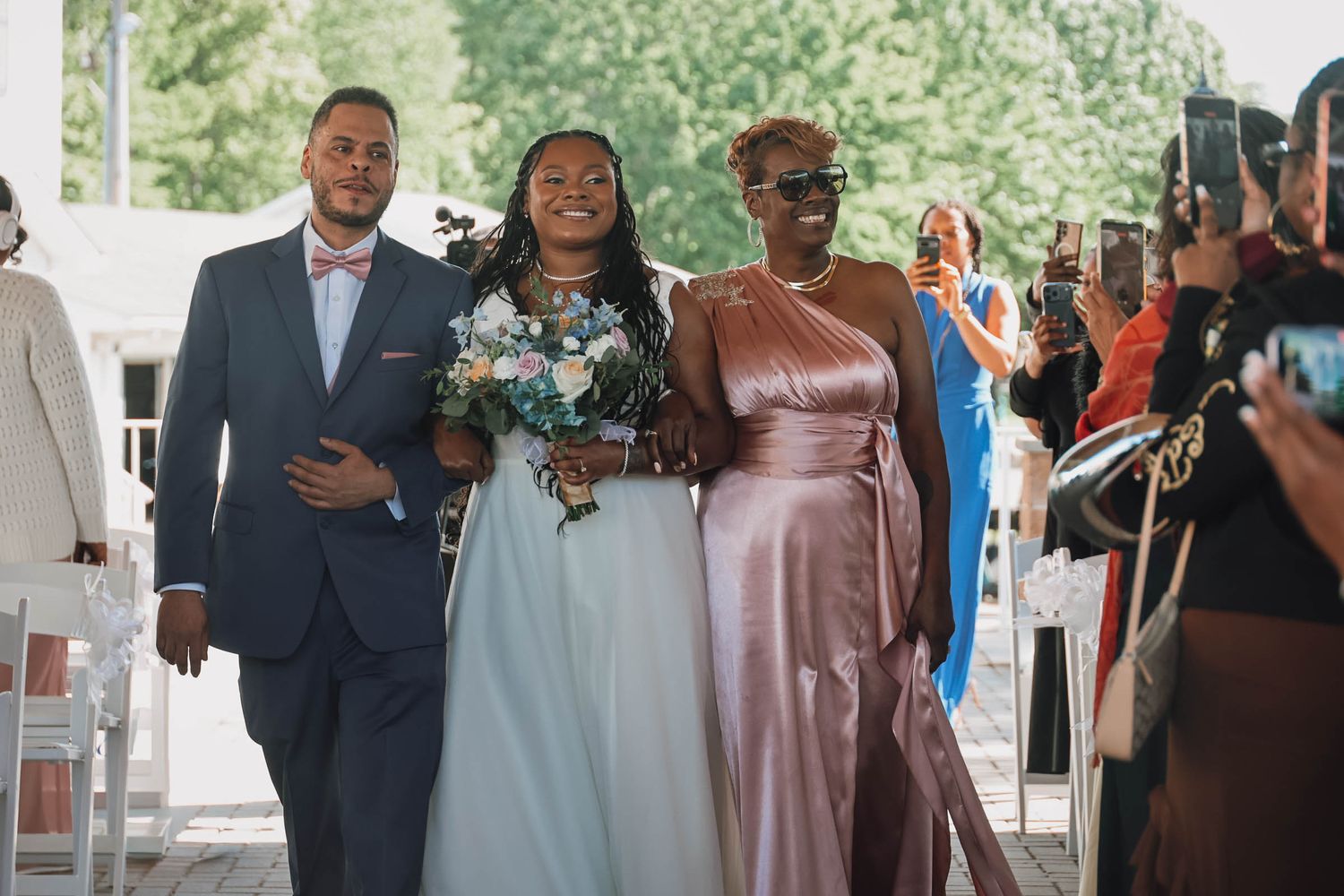 Bride walks down the aisle escorted by two family members at an outdoor wedding ceremony.