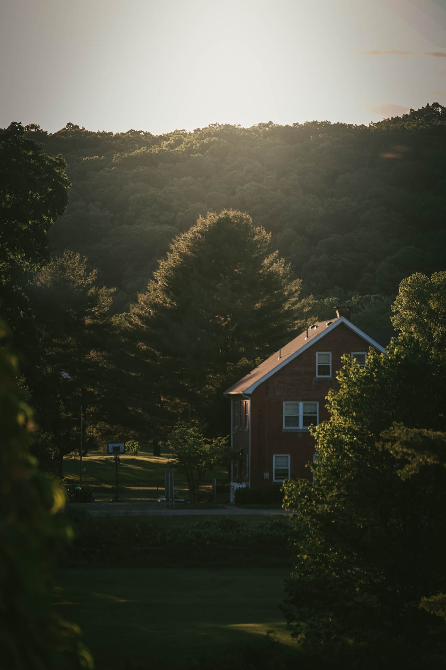 Red brick farmhouse nestled among lush green trees on a hillside at golden hour sunset in rural countryside.