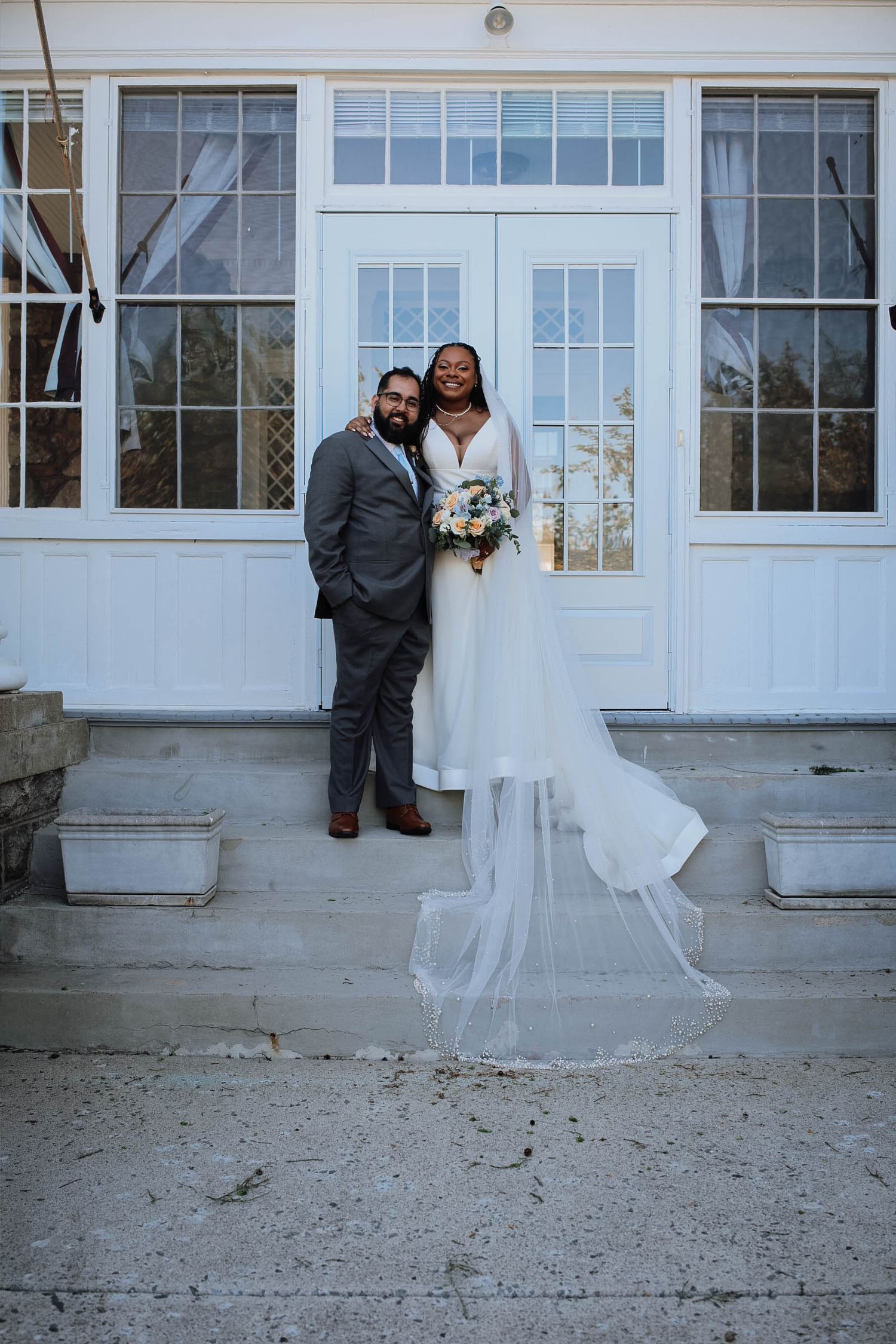 A bride in a flowing white gown with veil and groom in gray suit pose on steps before white French doors.