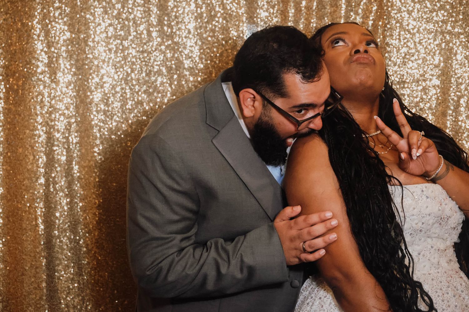 Couple posing playfully in a photo booth with gold sequin backdrop, man kissing woman's cheek at a wedding.