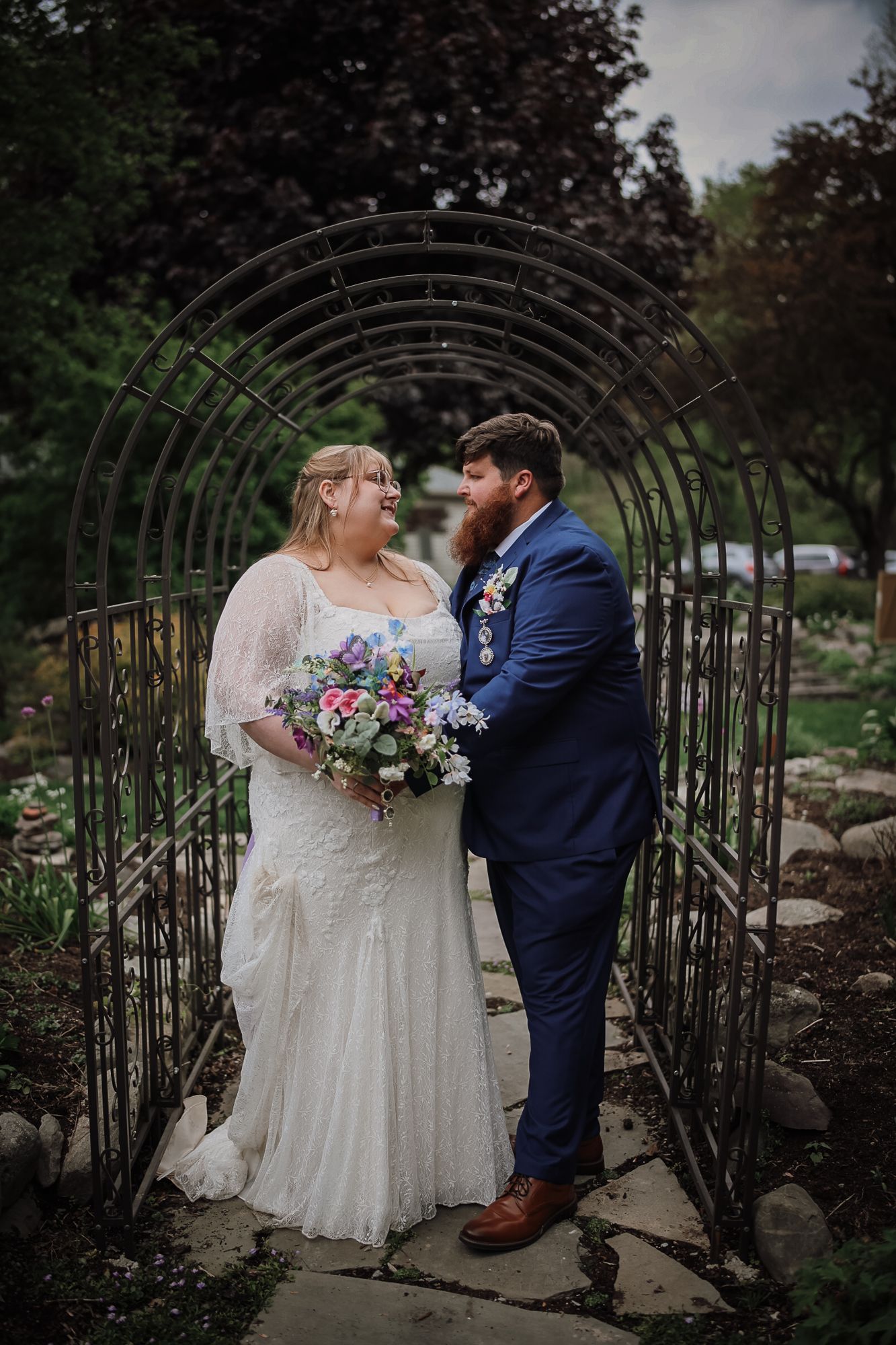 Bride and groom share a romantic moment under a metal arch in a garden, bride holding colorful bouquet, groom in navy suit.