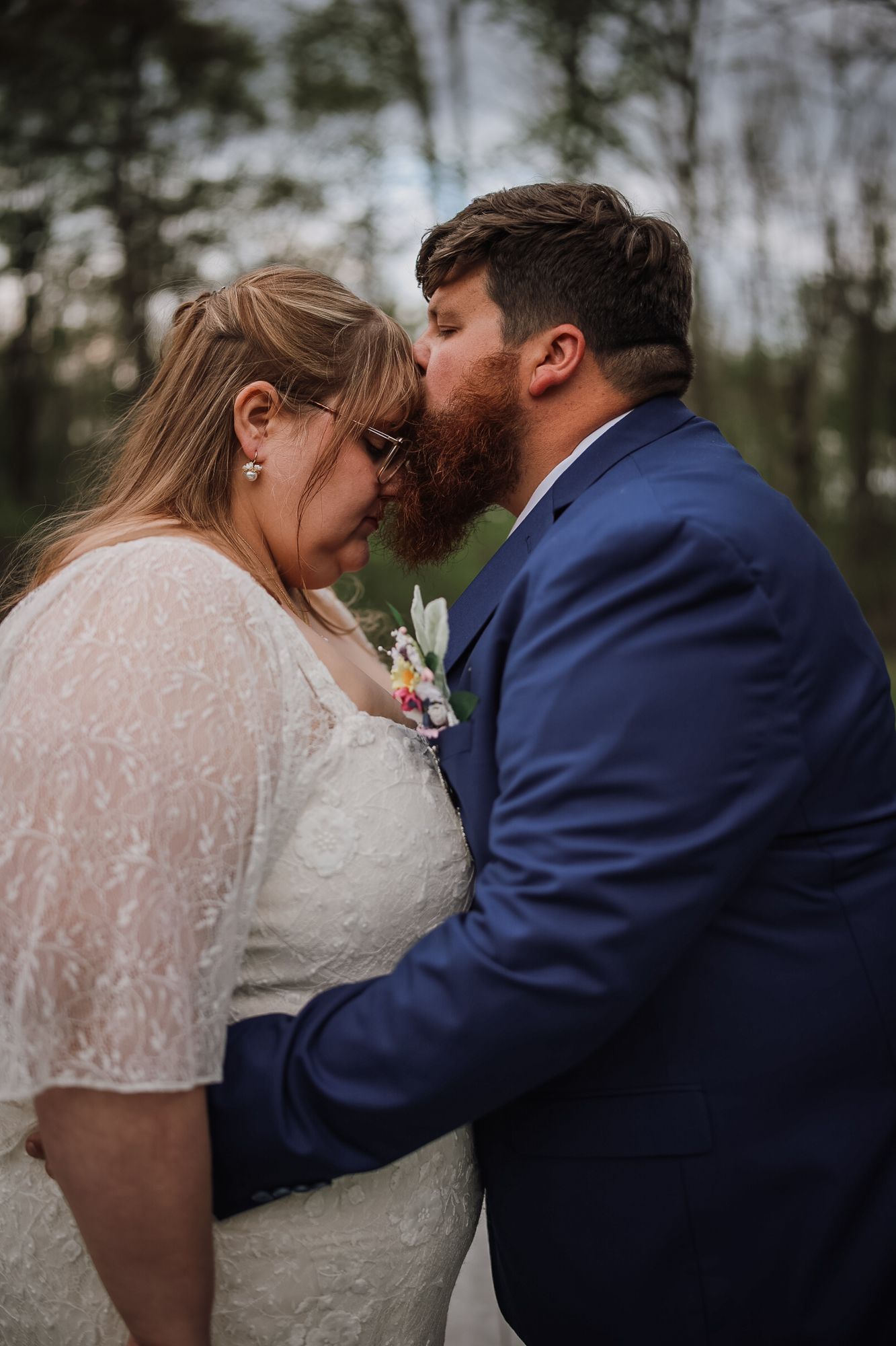 A bearded groom in a blue suit kisses his bride in a white lace dress on the forehead outdoors.