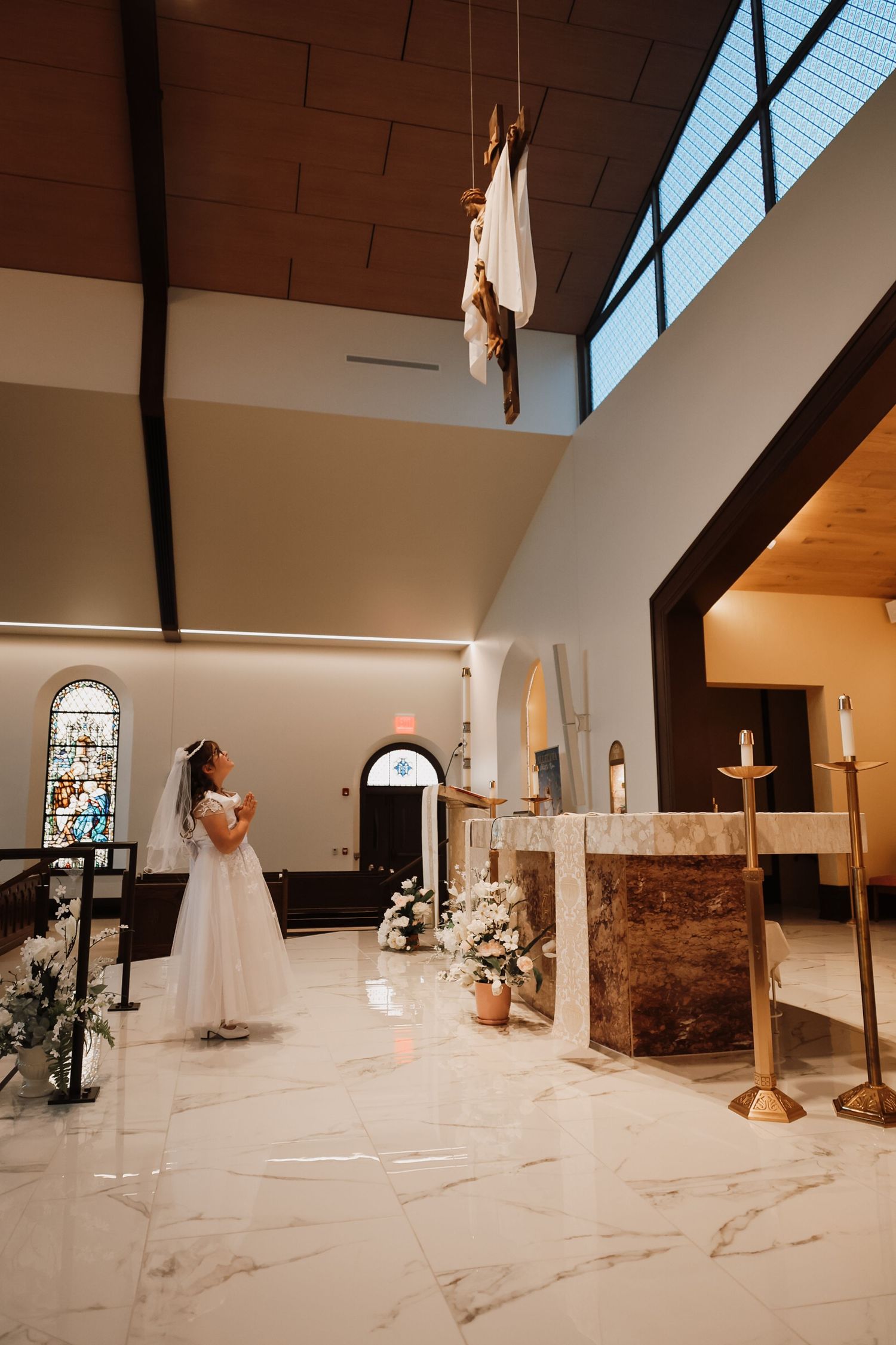 Young girl in white dress during First Communion ceremony at a Catholic church altar with crucifix above.