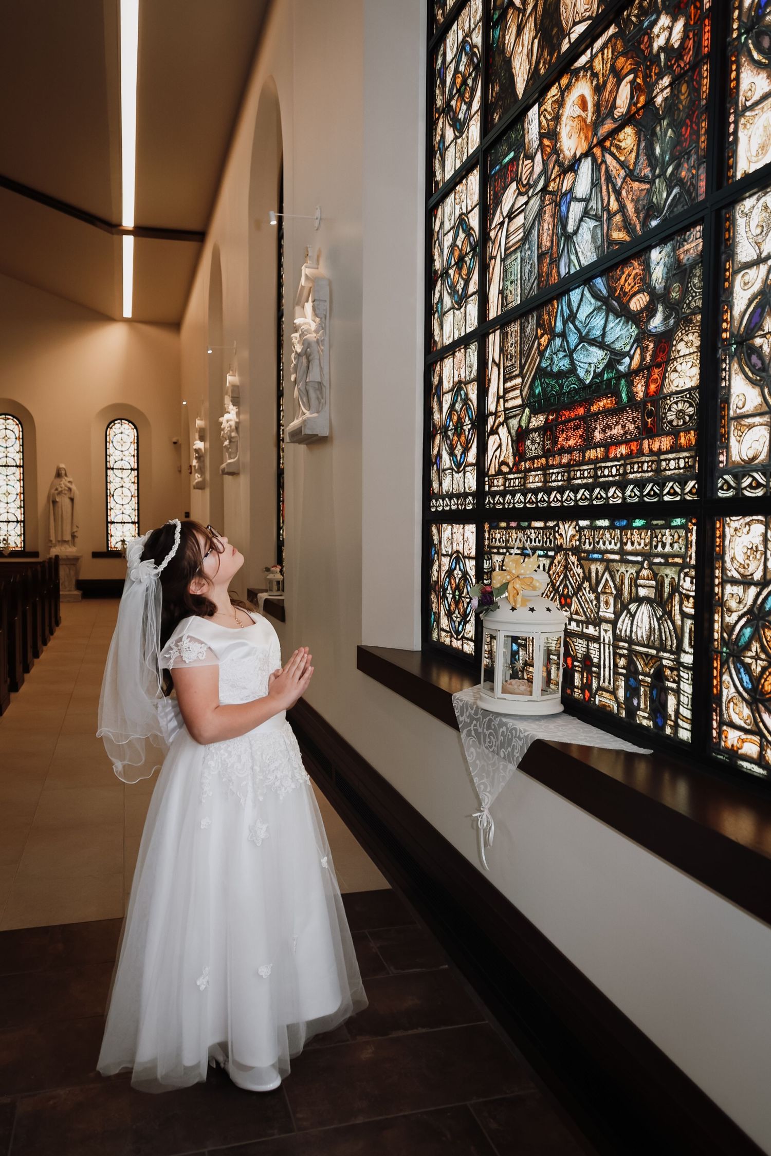 Young girl in white First Communion dress and veil praying before a colorful stained glass window in a church.