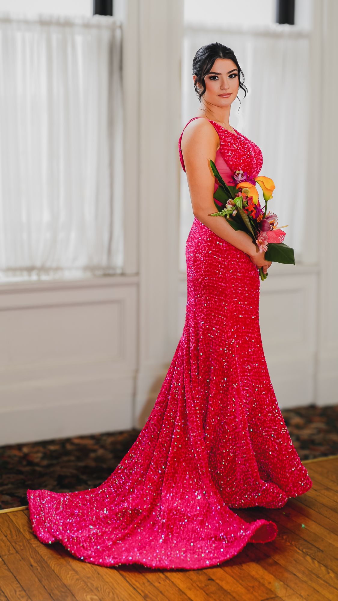Woman in a sparkling hot pink sequin mermaid gown holding a colorful bouquet, posing elegantly indoors.