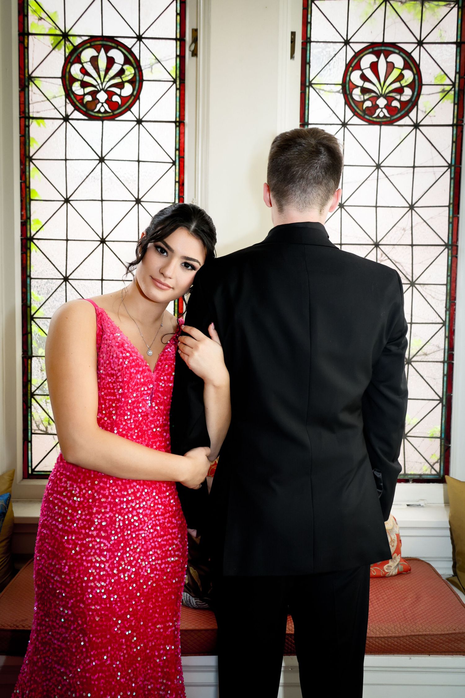 Young couple in formal wear posing by stained glass window, woman in red sequin gown, man in black suit.
