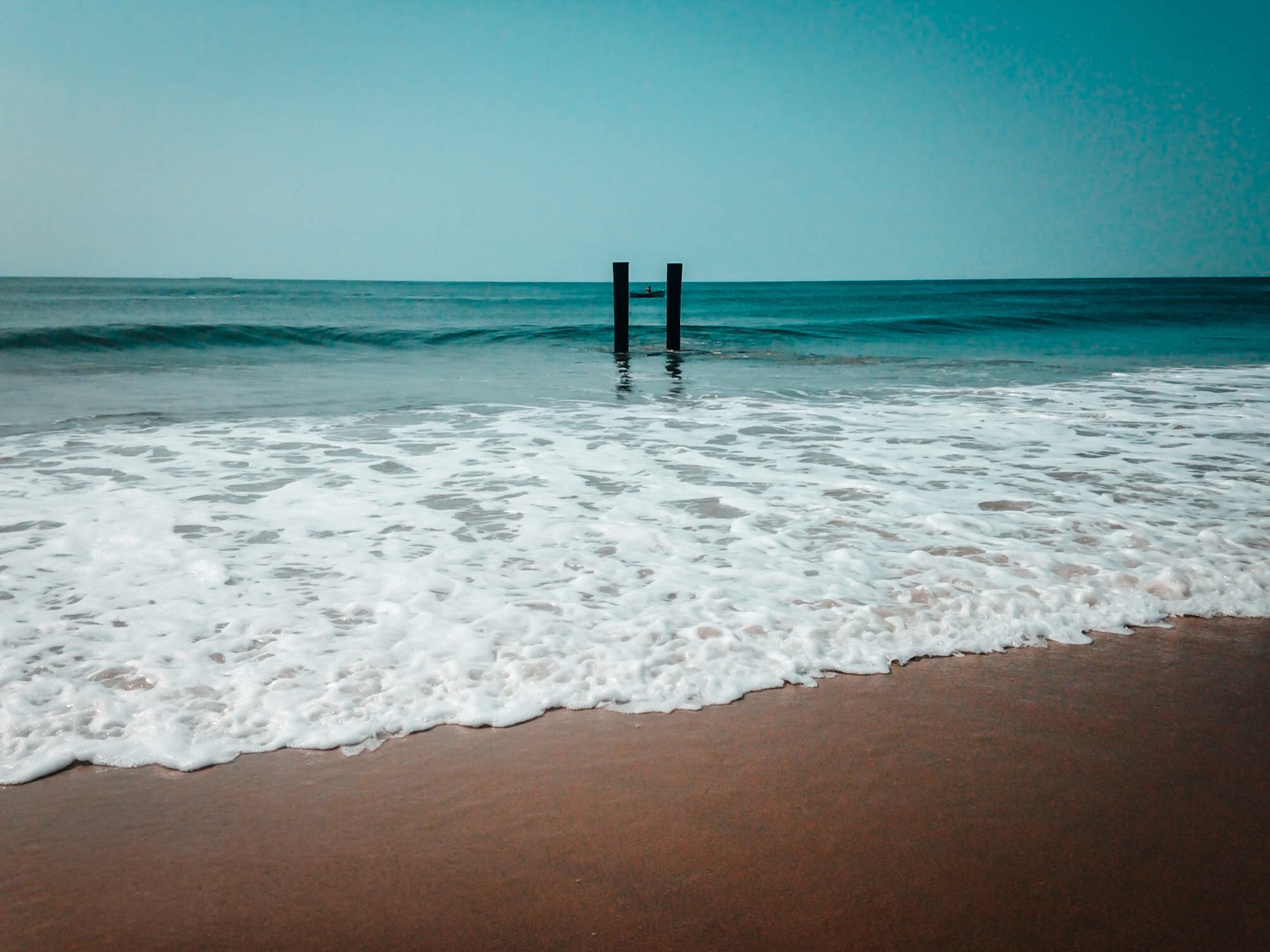 Calm ocean waves washing onto a sandy beach with two wooden posts standing in the shallow turquoise water.