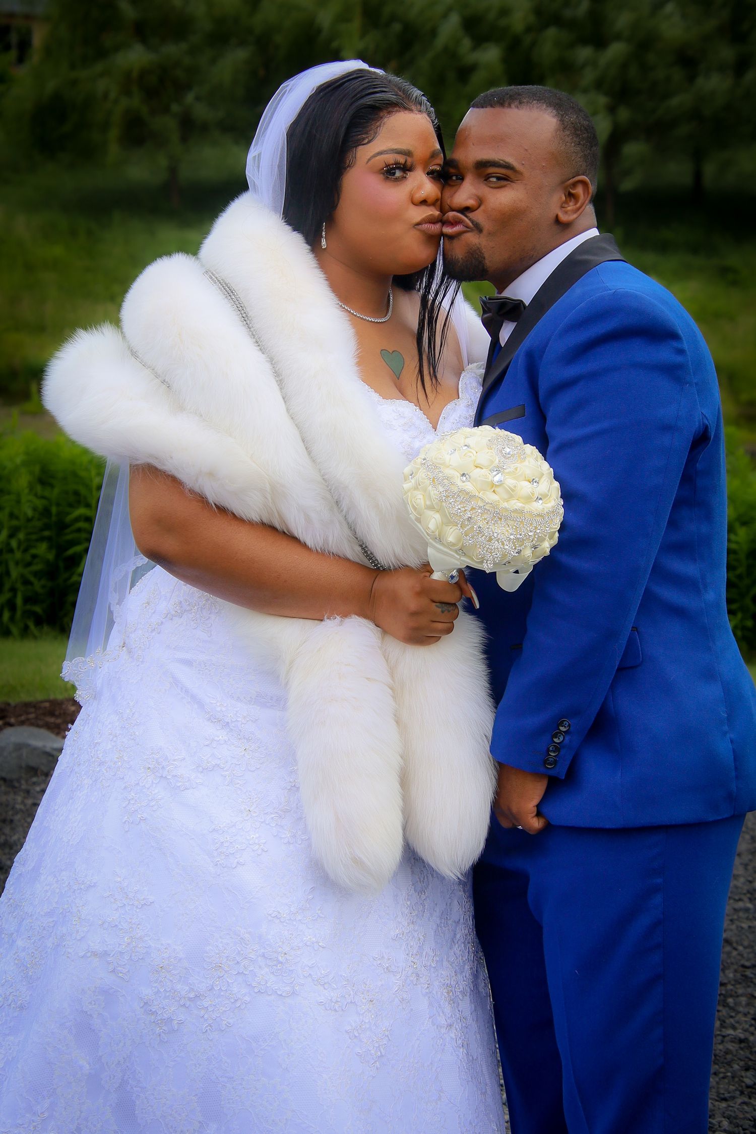 A romantic wedding couple in a blue suit and white dress with fur shawl share intimate moments in an outdoor setting.