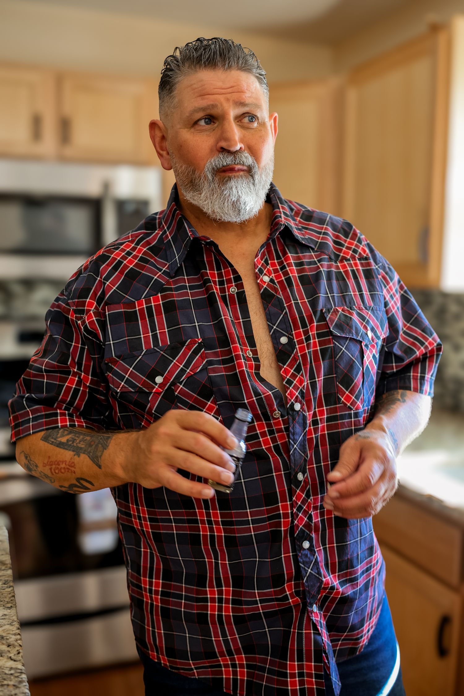 A person in a red plaid flannel shirt with tattoos and a gray beard stands in a kitchen setting.