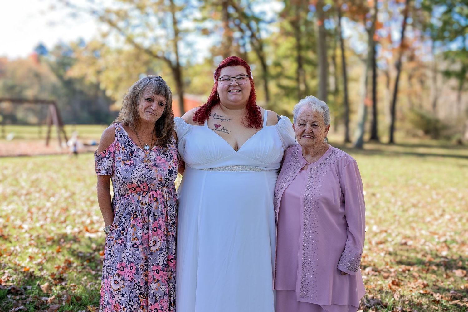 Three women stand together in a sunny autumn park with fallen leaves on the ground.