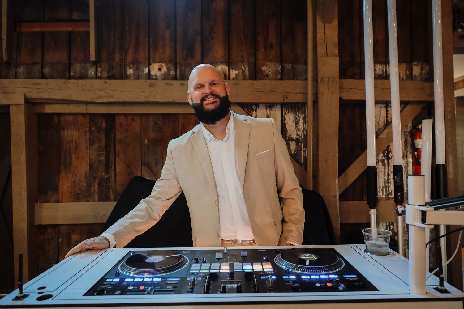 DJ stands behind professional mixing equipment at a rustic wooden venue wearing a tan blazer and white shirt.