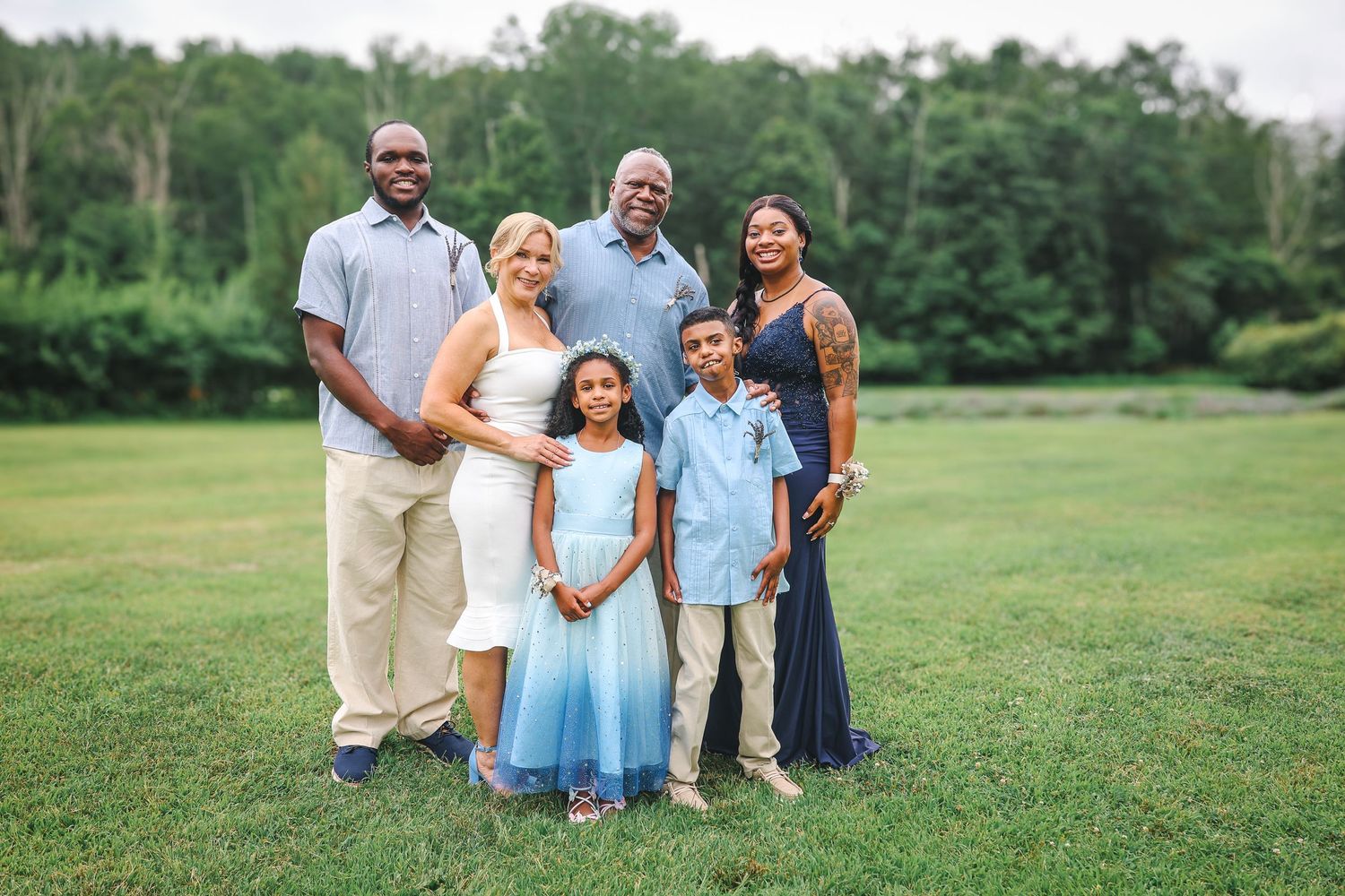 A family wearing coordinated blue and white outfits poses together on a grassy field during a summer photo session.