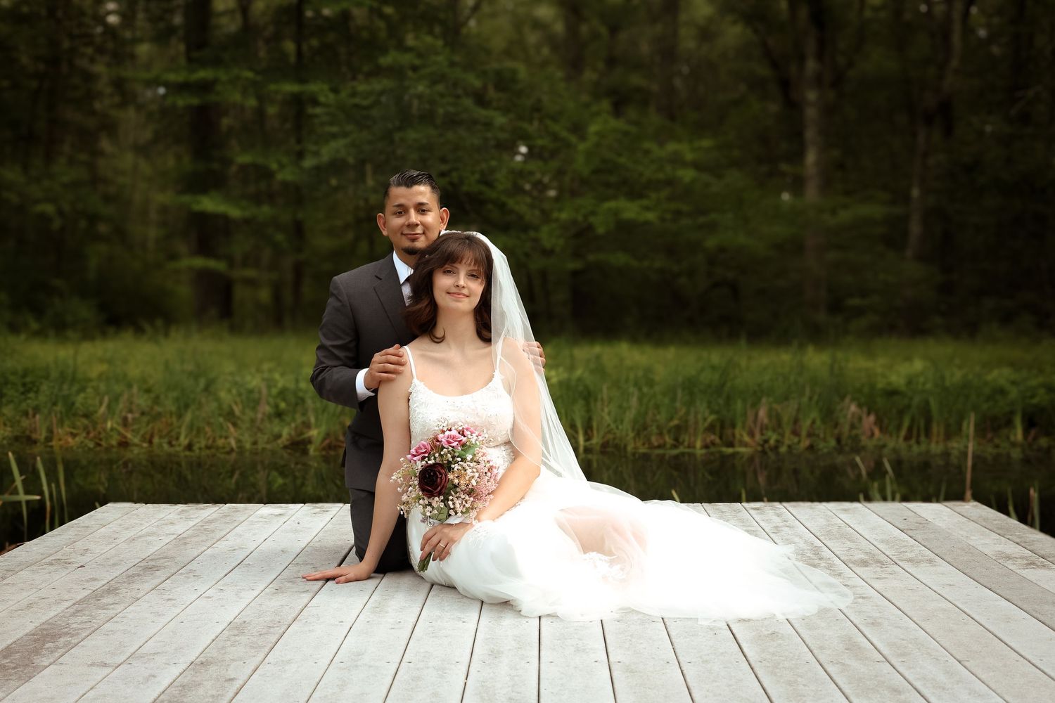 A couple poses for romantic wedding photos on a rustic wooden dock surrounded by lush greenery and trees.
