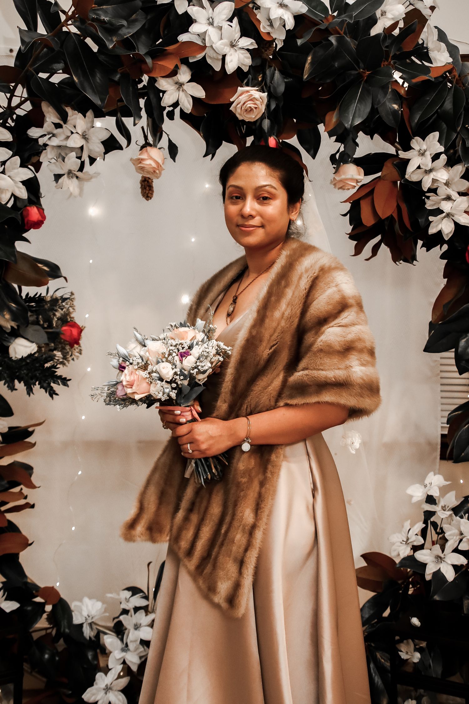 A person wearing a beige dress and brown fur stole poses elegantly against a backdrop of white flowers and greenery.