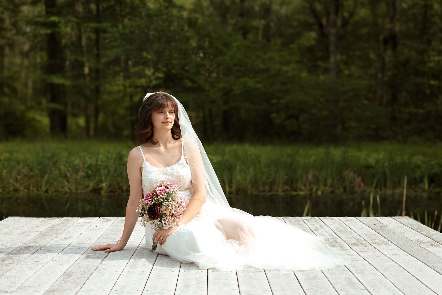 A bride in a white wedding dress sits gracefully on a wooden deck surrounded by lush greenery.