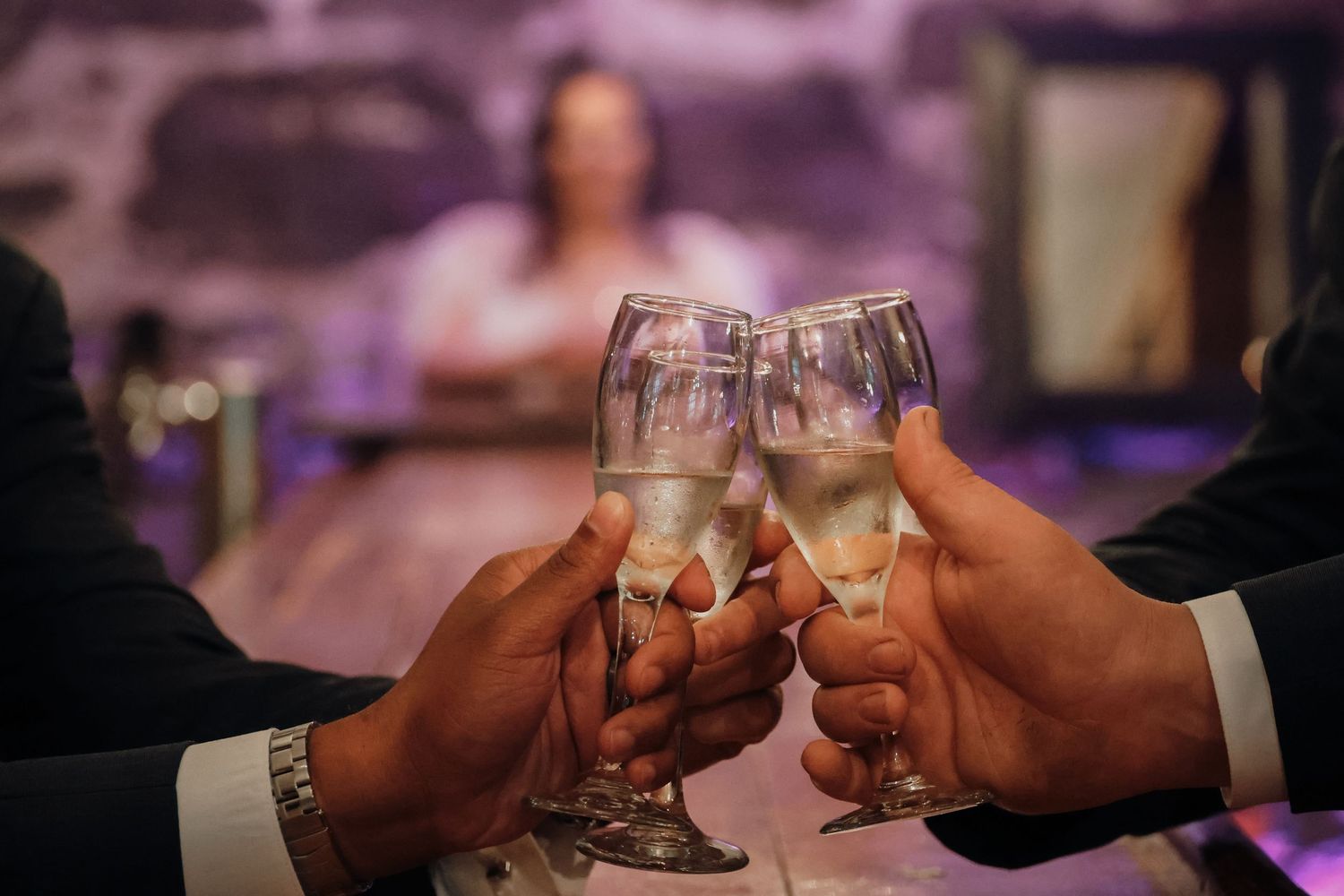 Hands clinking champagne glasses in celebration at an elegant event with purple ambient lighting in the background.
