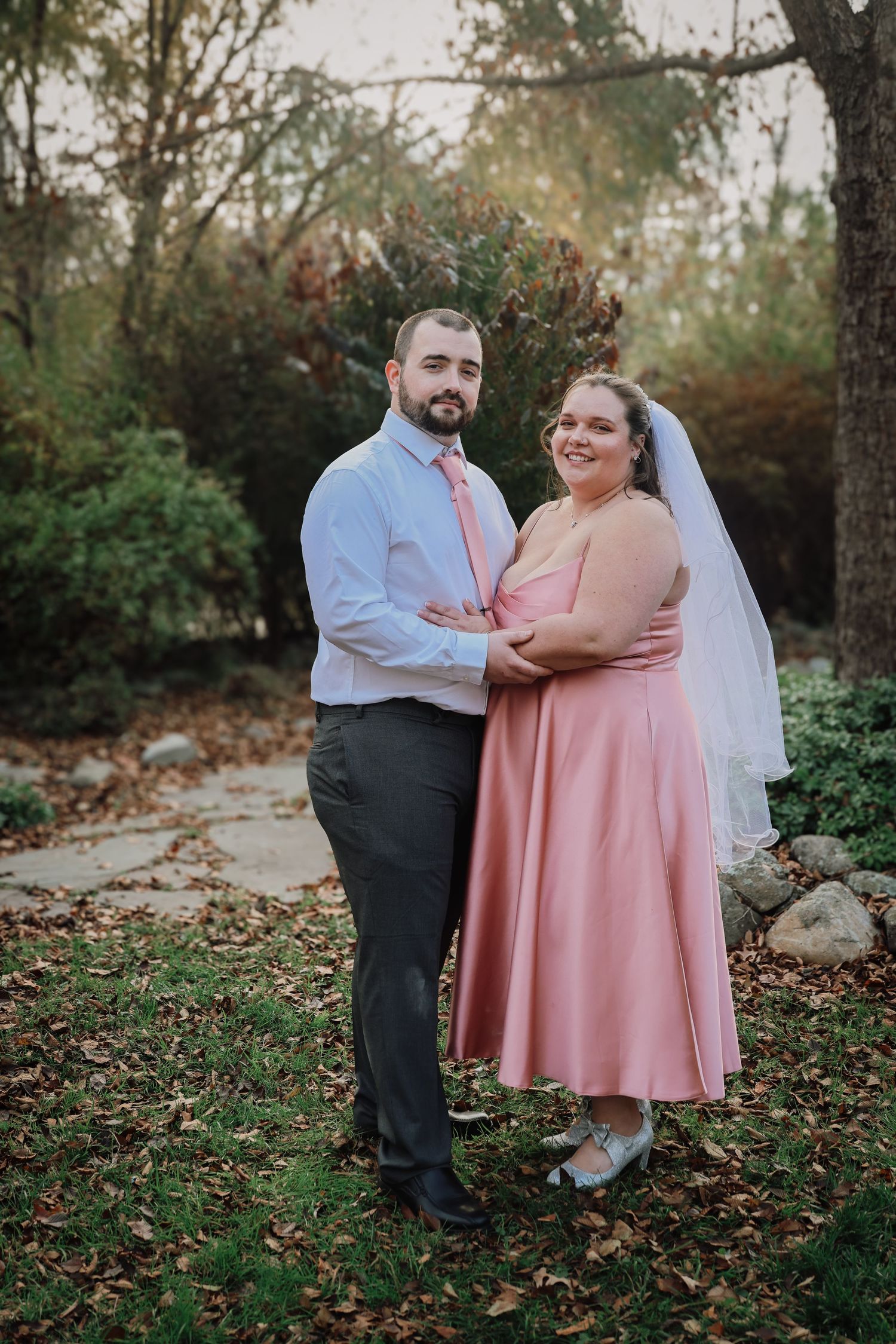 A couple in wedding attire shares a romantic moment outdoors during autumn with fallen leaves on the ground.