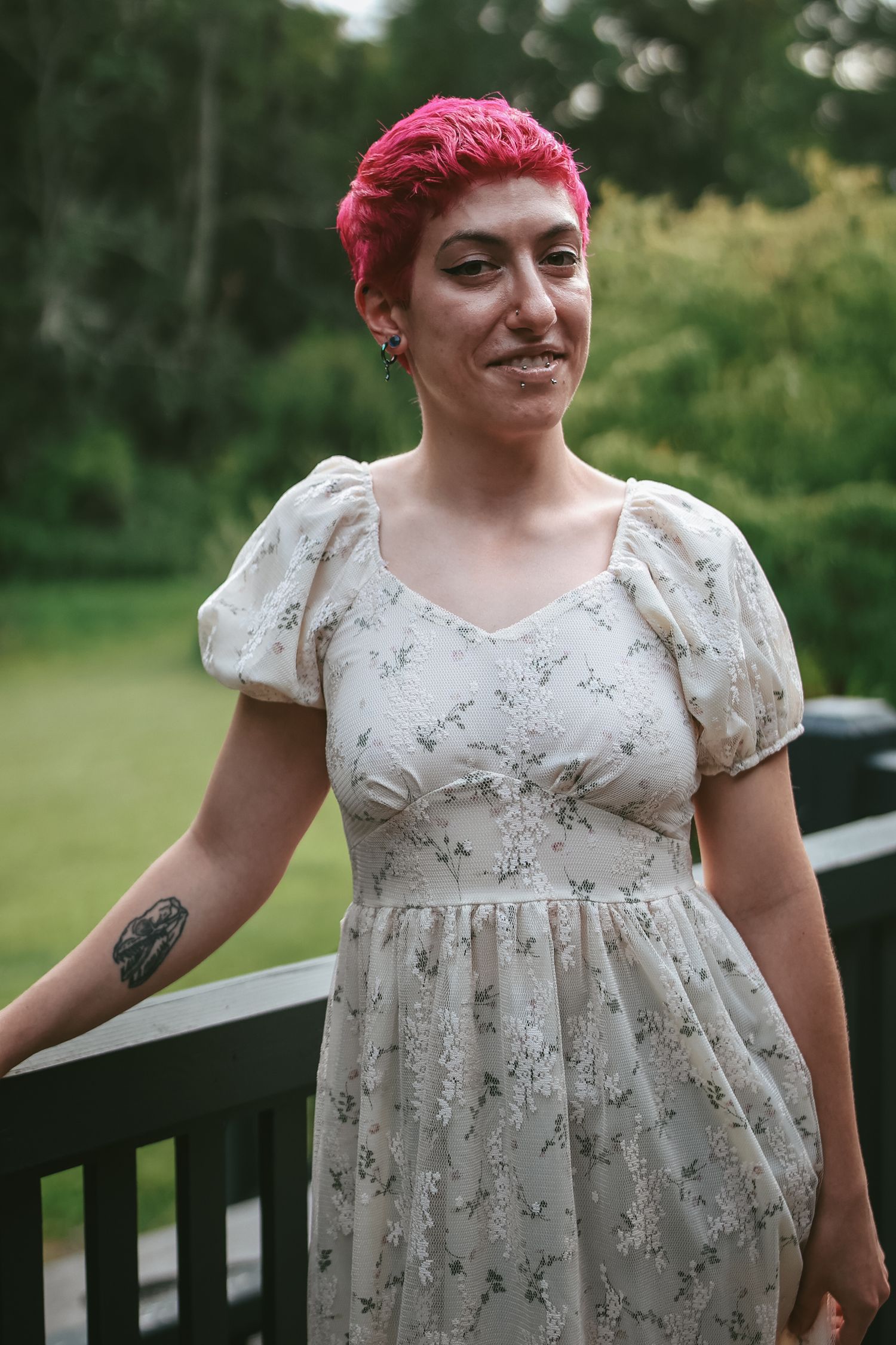 A person with vibrant pink hair wearing a white floral dress poses outdoors on a wooden deck.