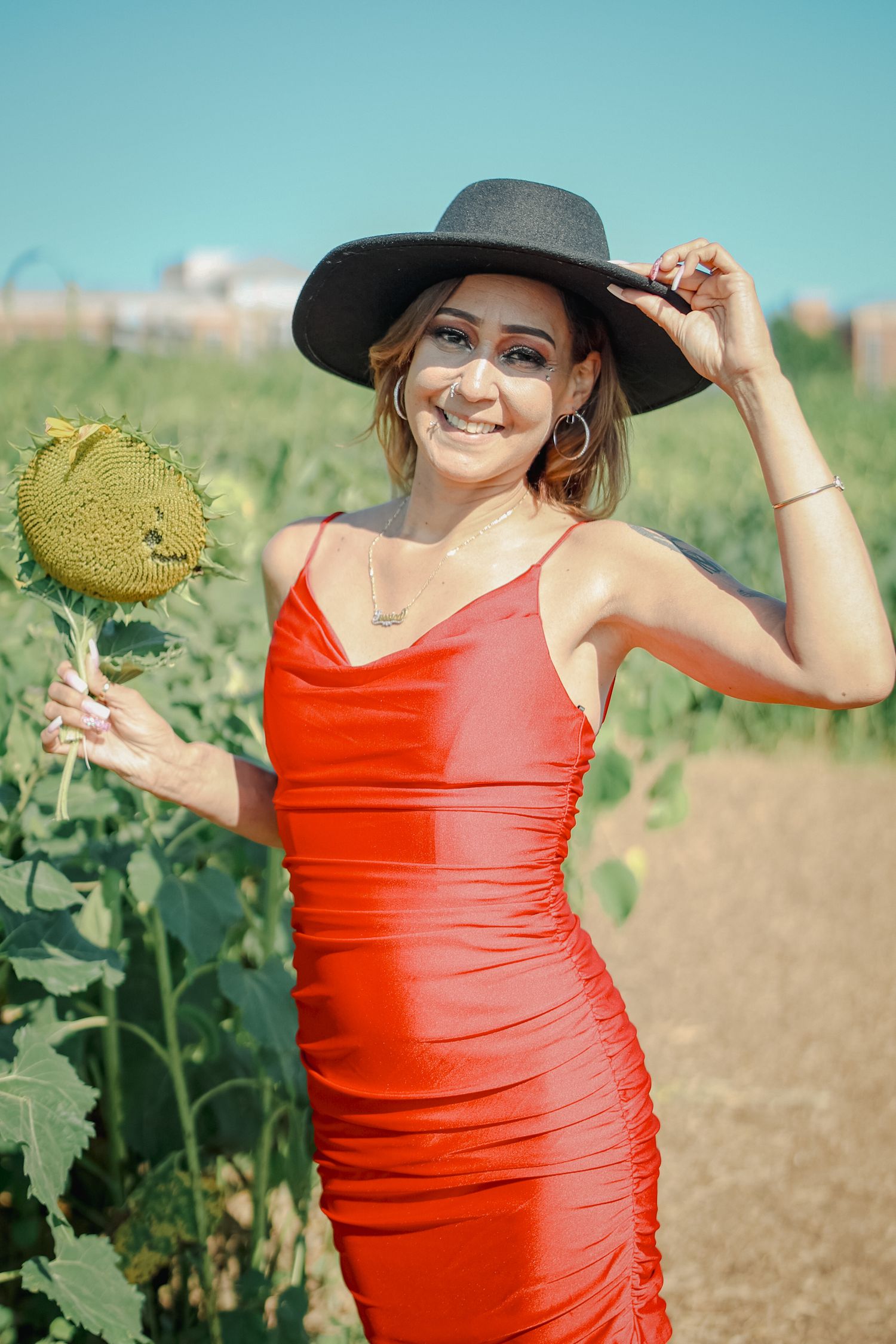 A person in a bright red dress and sun hat poses playfully in a sunflower field on a sunny summer day.