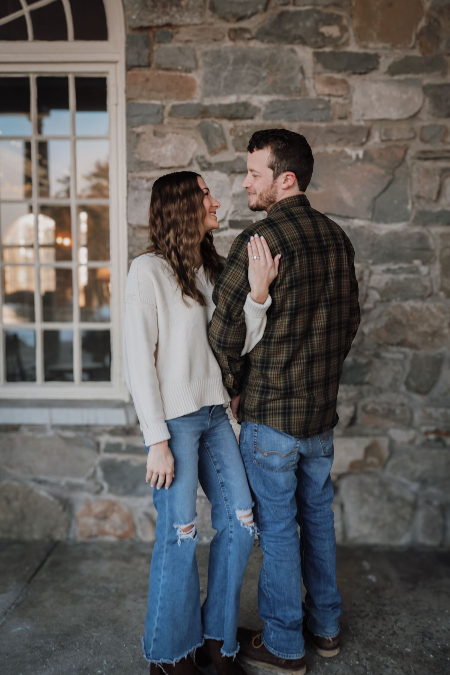 A couple shares a romantic moment against a rustic stone wall with windows on a bright day.