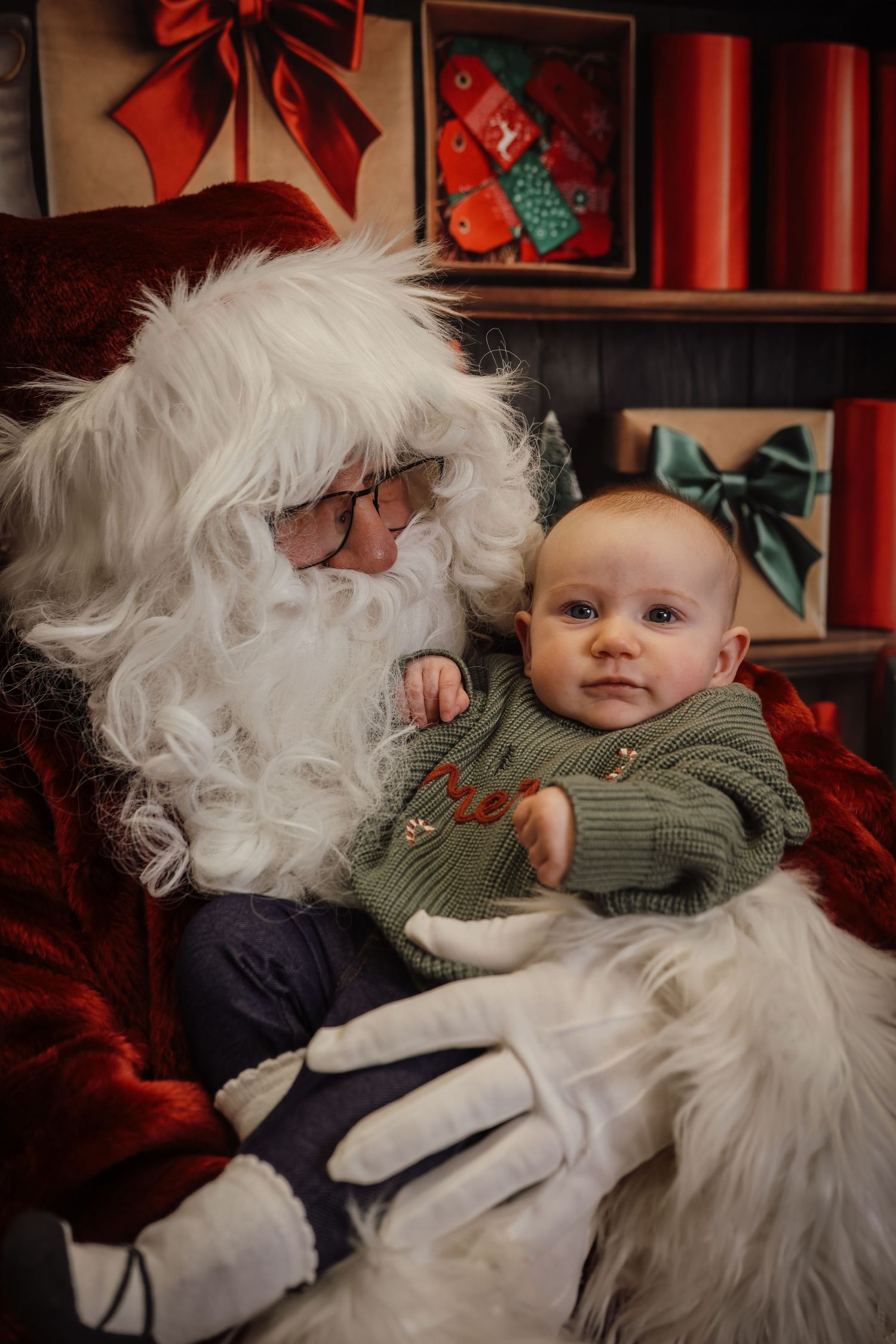 A baby in a green sweater sits with Santa Claus in a festive holiday setting with red bows and decorations.