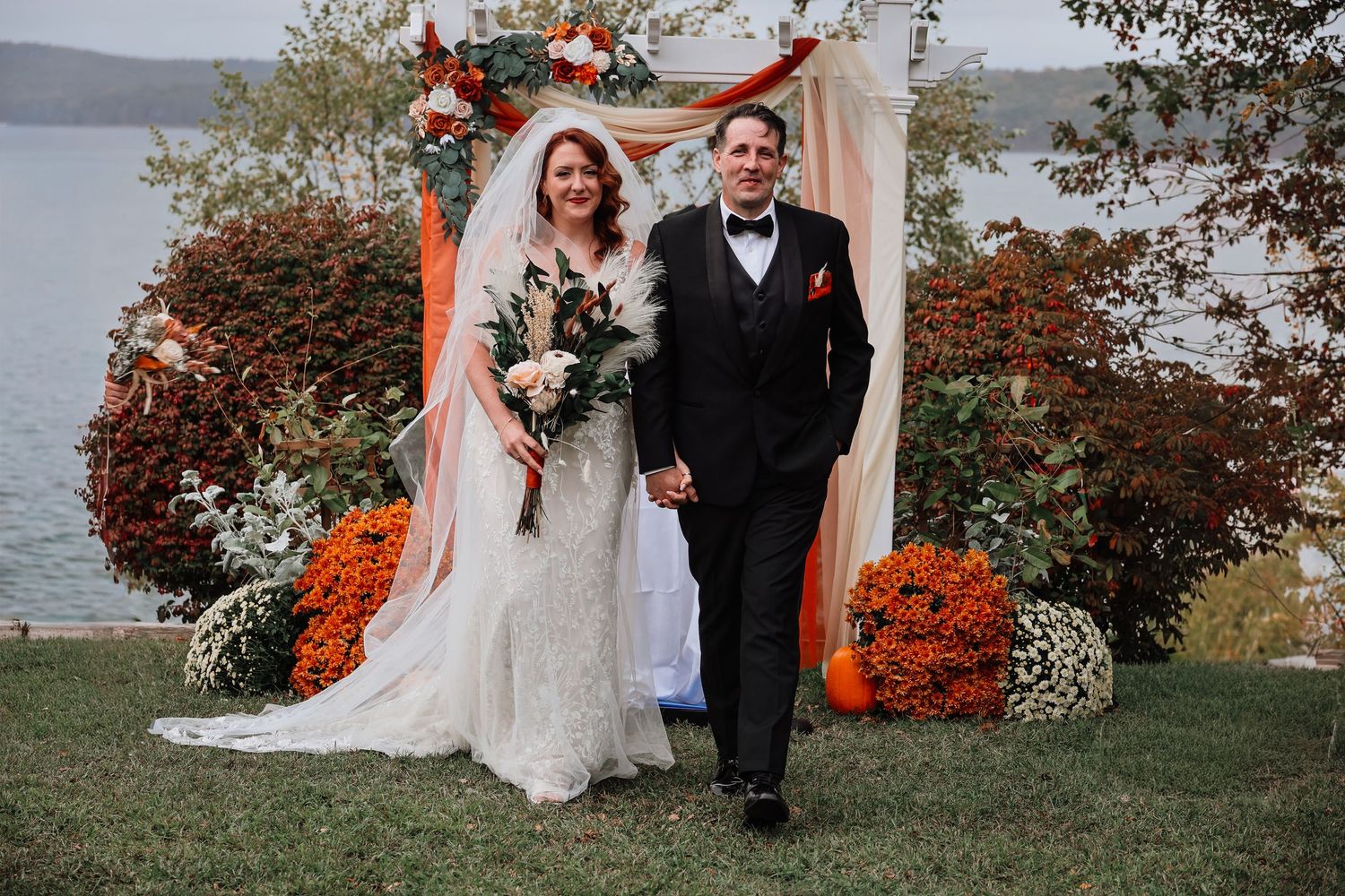 A couple poses at their autumn lakeside wedding with vibrant orange fall foliage and waterfront views in the background.