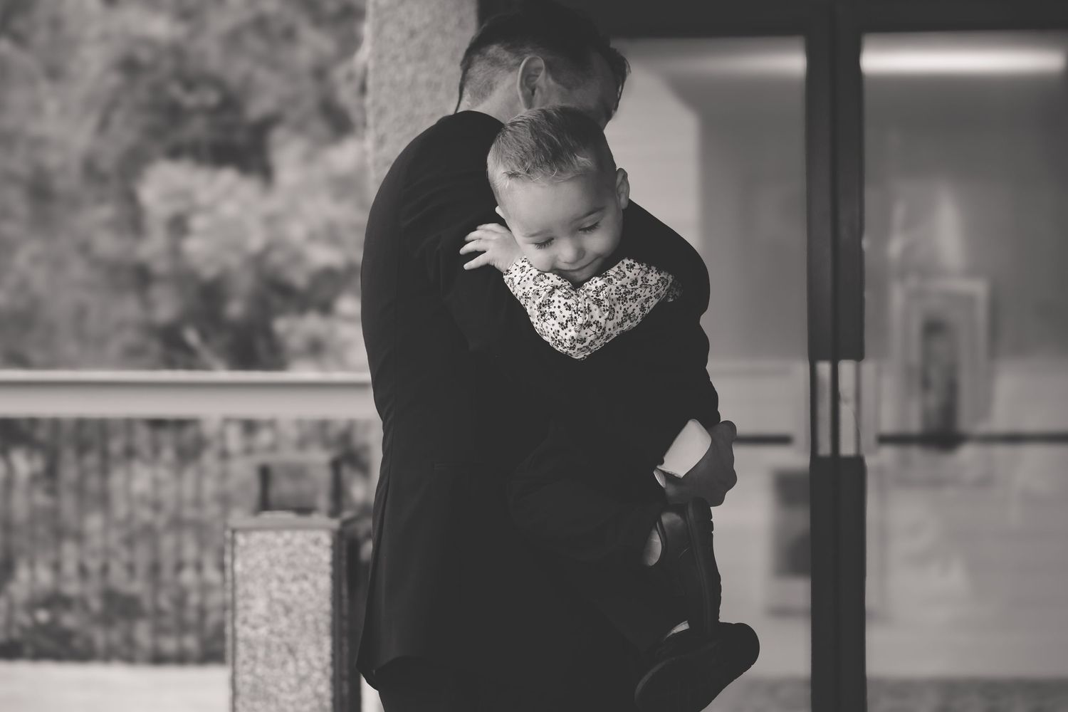 A tender black and white photograph showing a loving embrace on a porch with soft natural lighting.