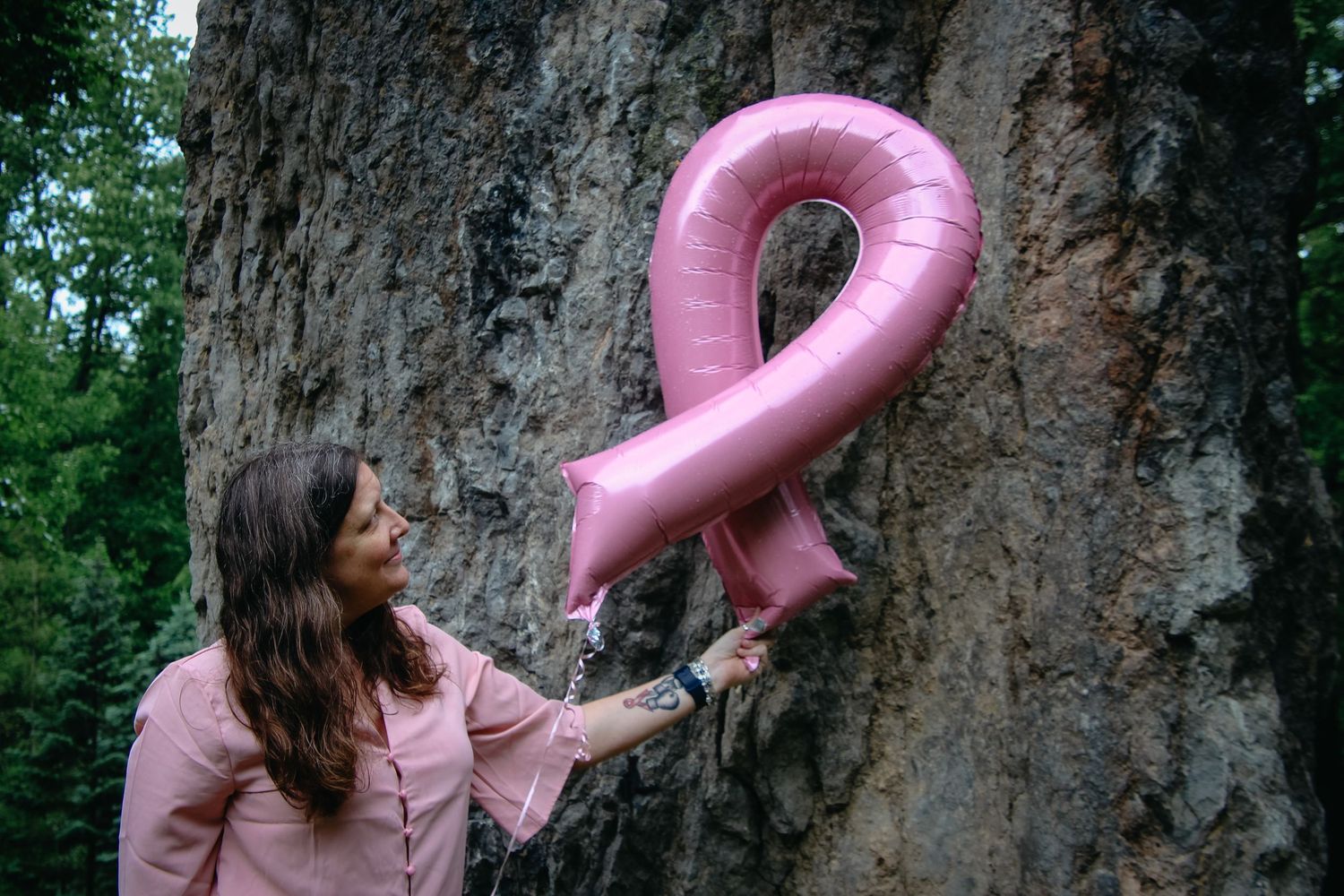 A pink balloon flamingo appears to float against a tree trunk in a forest setting.