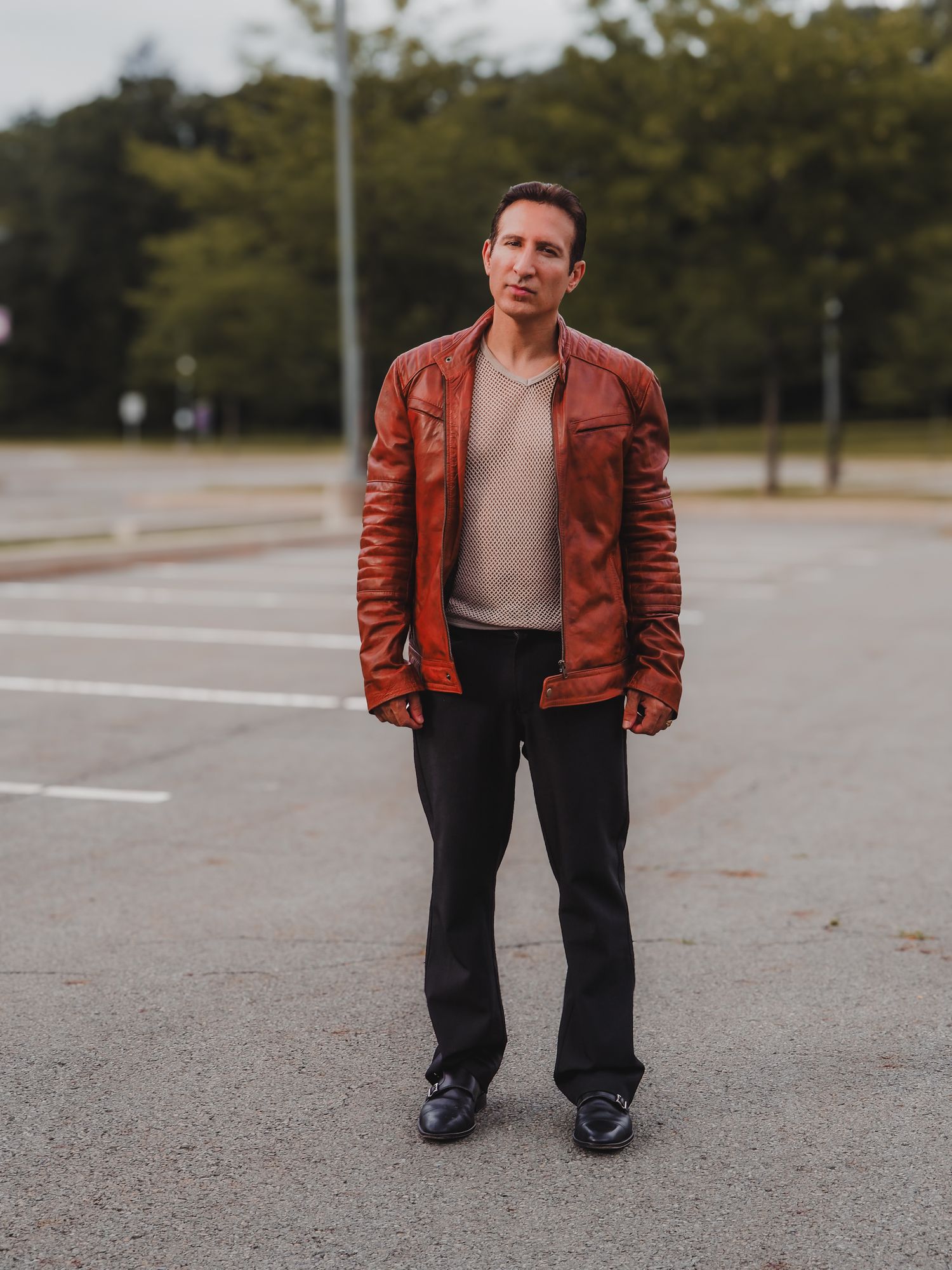 A person in a brown leather jacket and gray sweater stands in an empty parking lot on an autumn day.