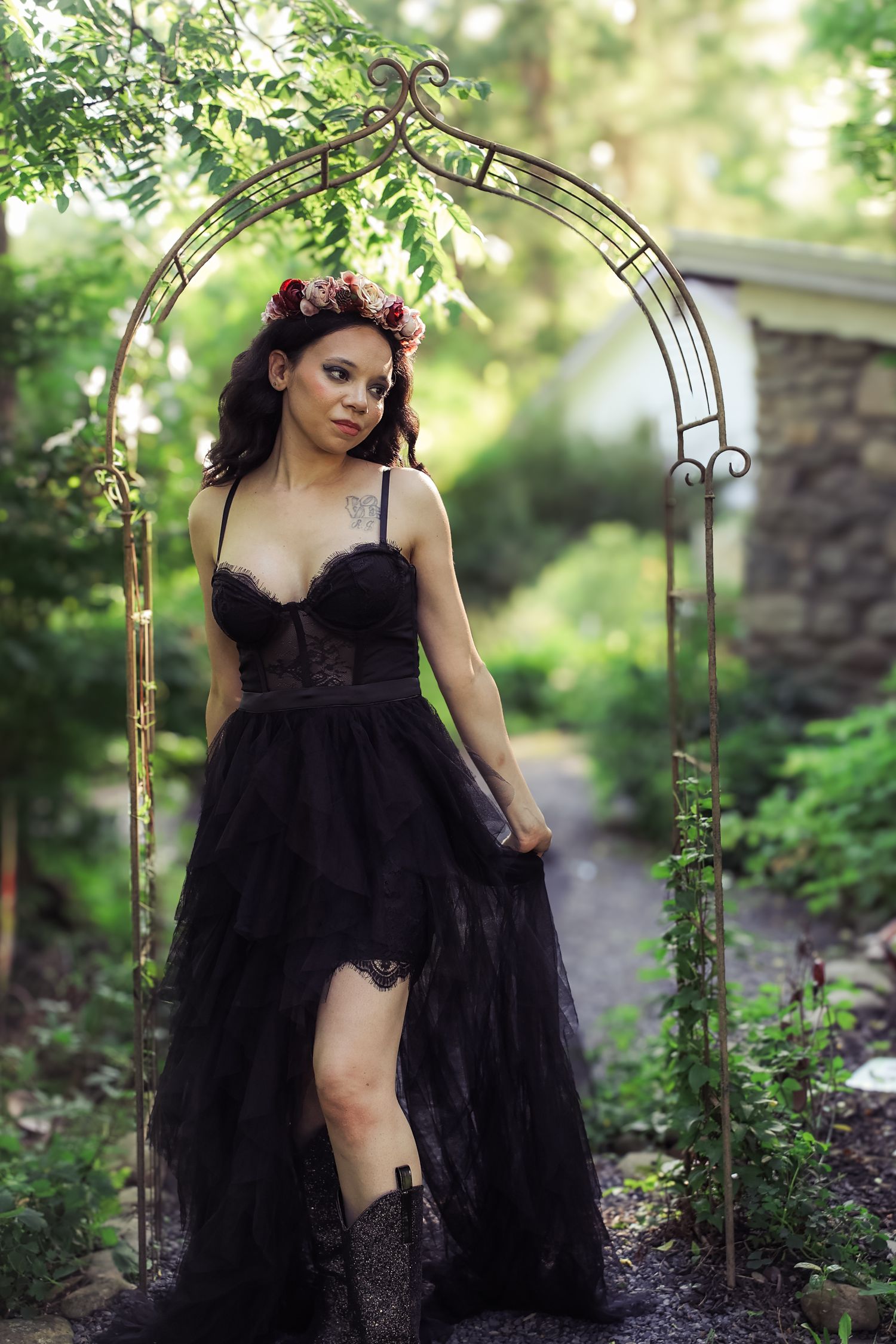 A figure in a flowing black tulle gown poses gracefully in a garden setting with stone walls and greenery.