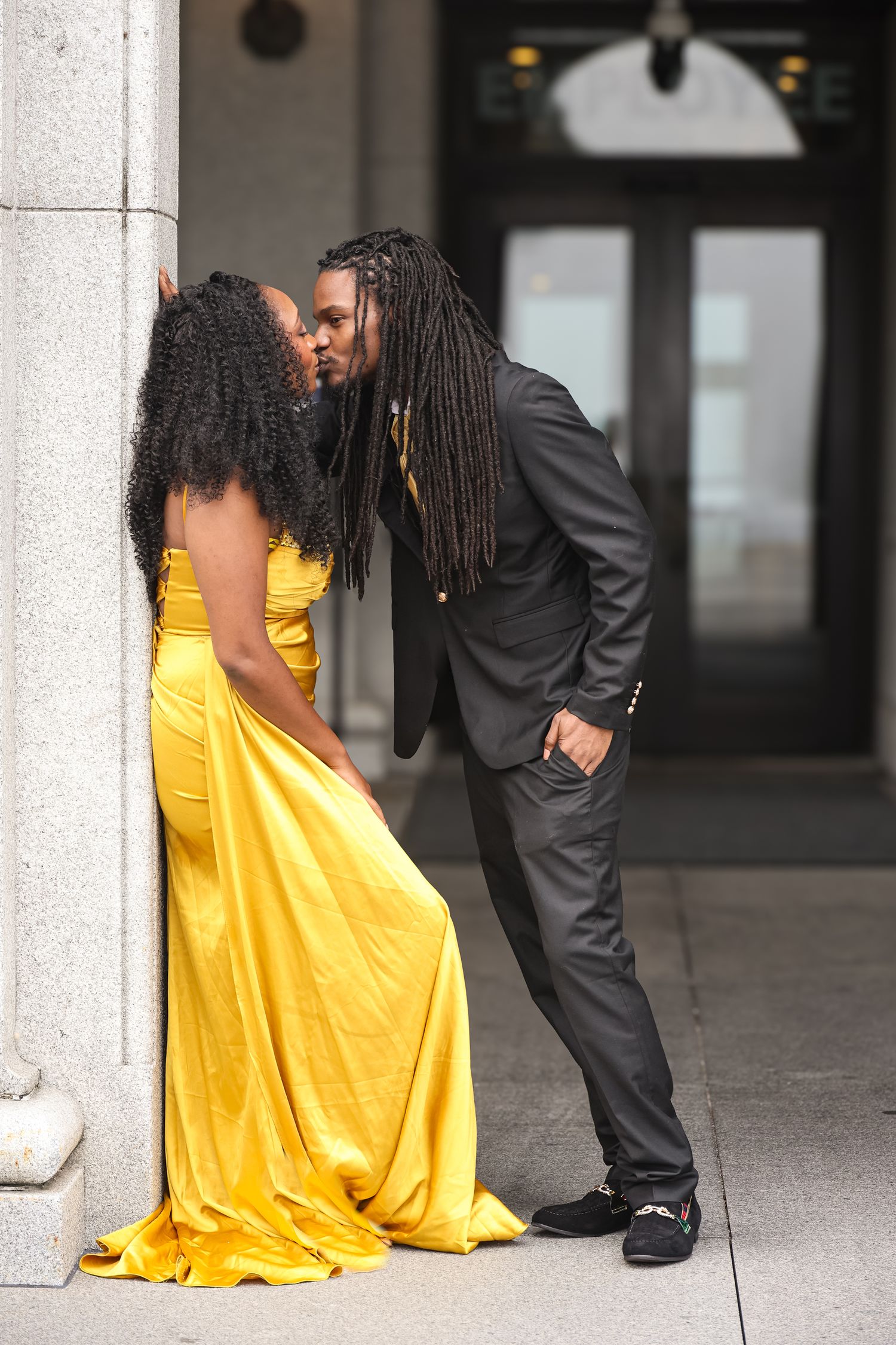 A person in a flowing yellow dress sits on steps while someone in a dark suit stands nearby in a building interior.