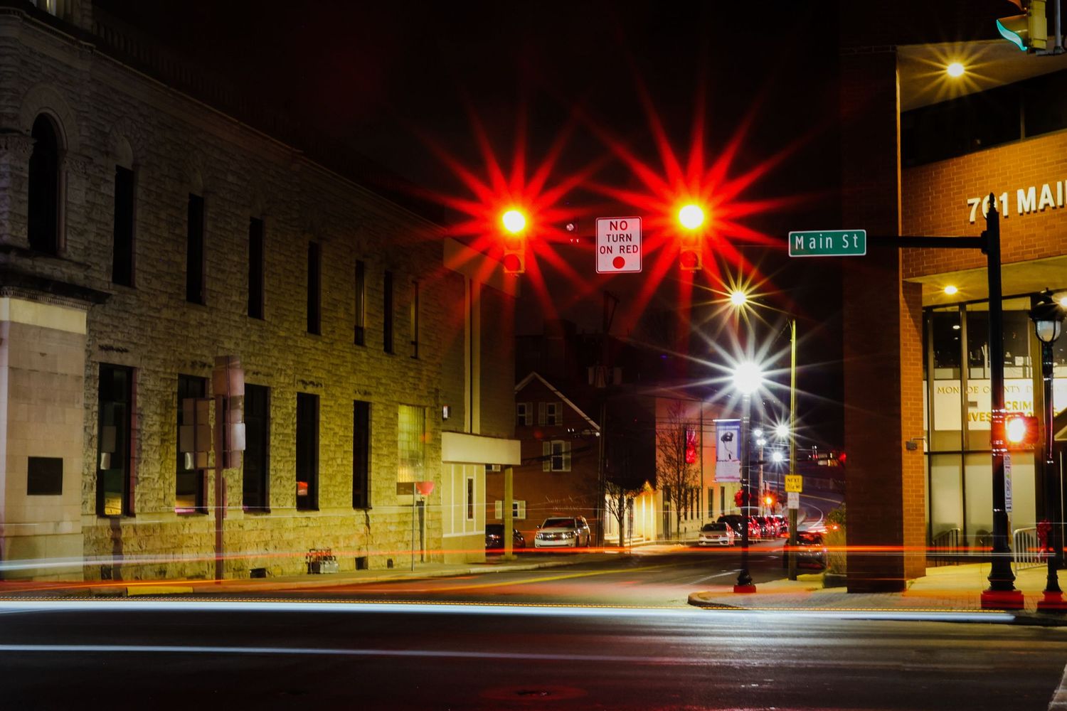 Red traffic lights illuminate a quiet urban street corner at night with brick buildings and storefronts.