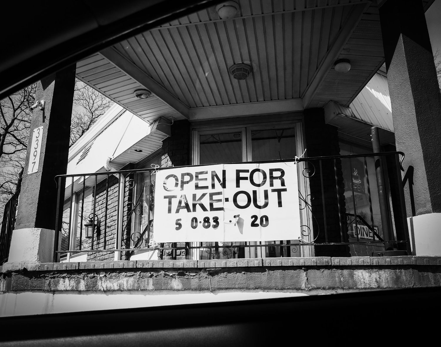 A black and white photograph showing a hand-written 'OPEN FOR TAKE-OUT' sign hanging on a building's front porch.