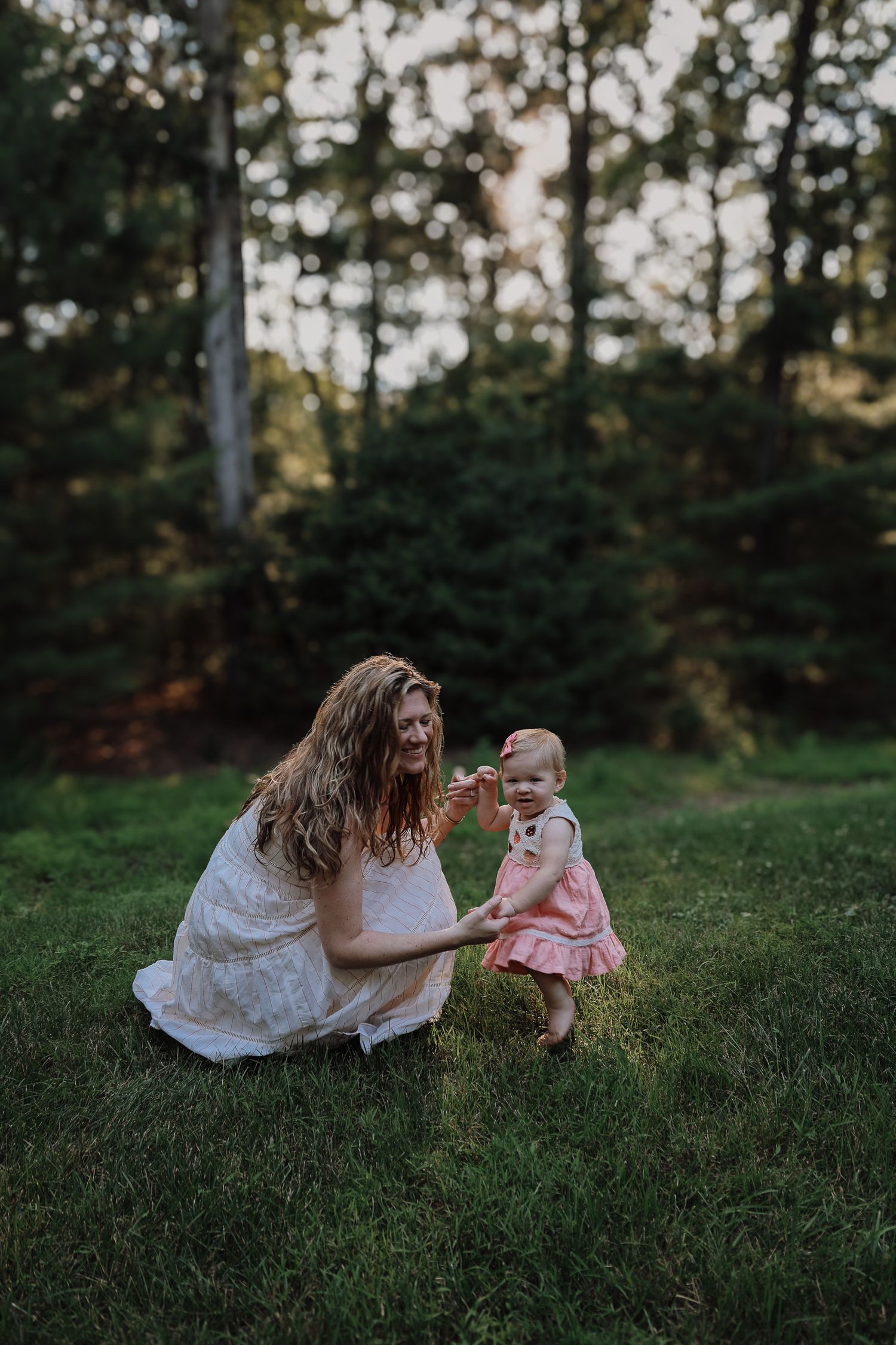 A woman in a white dress sits on grass with a baby wearing pink during an outdoor photo session.