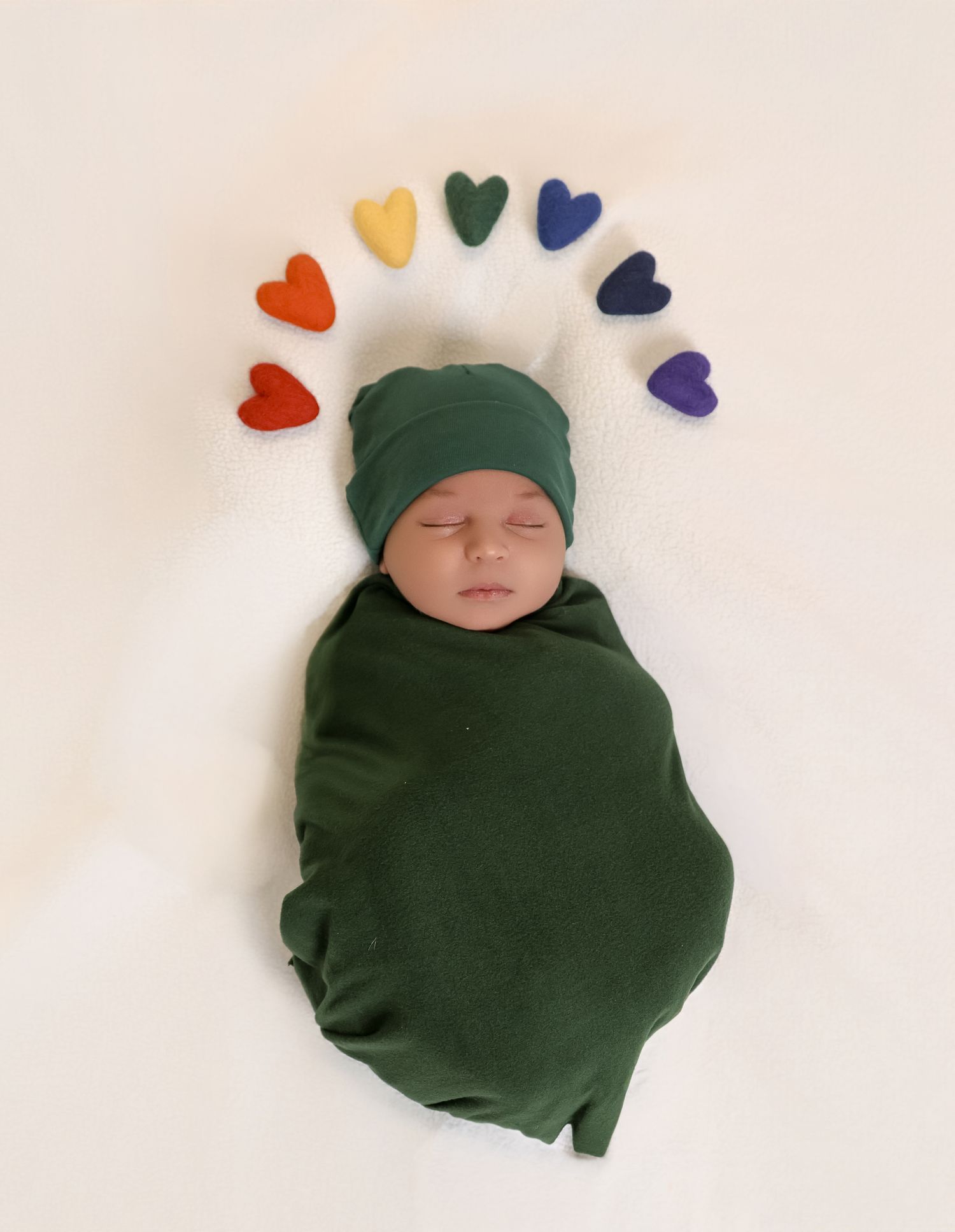 A sleeping newborn baby wrapped in dark green swaddle with colorful heart decorations on white background.