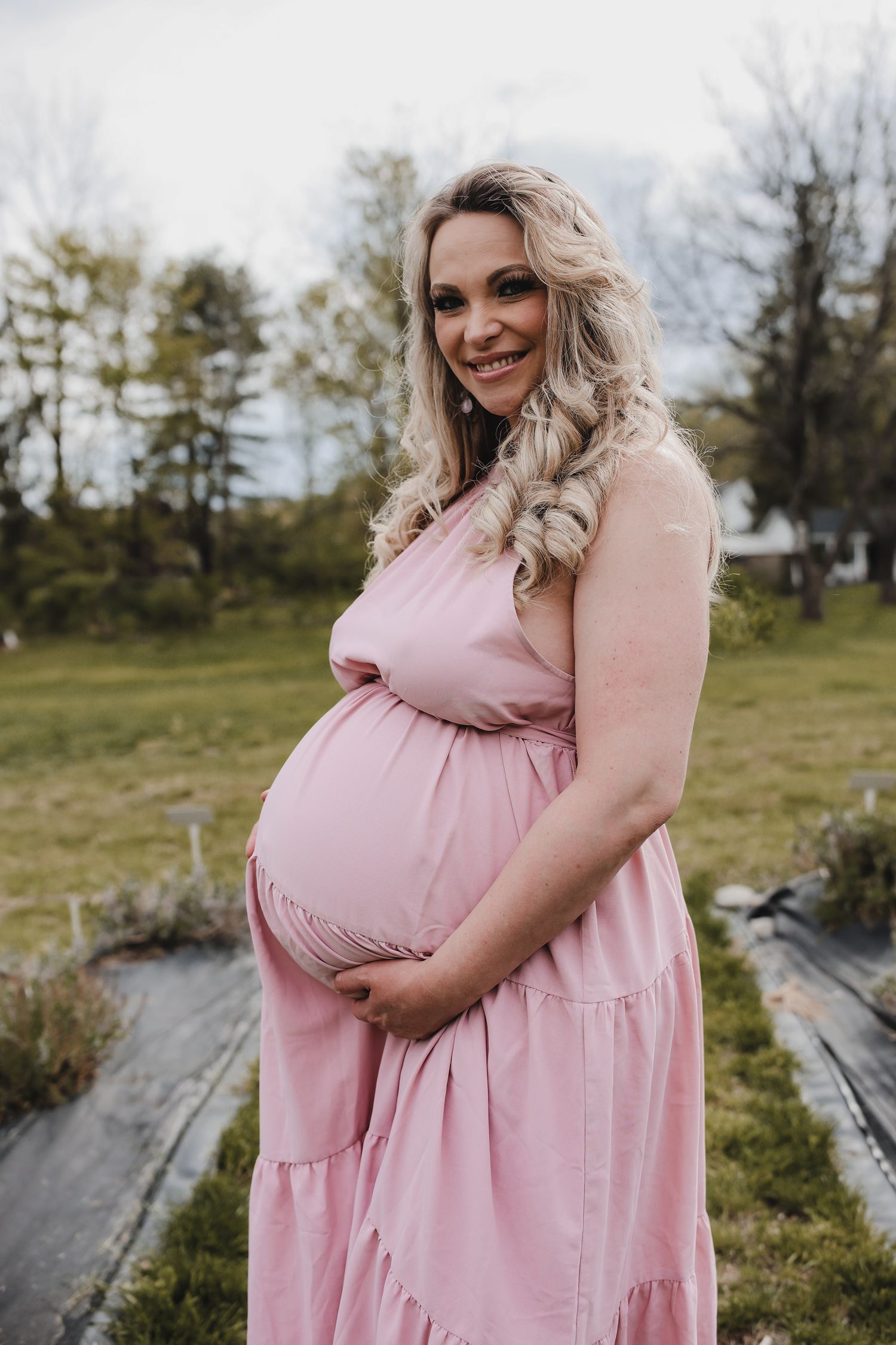 A person in a flowing pink maternity dress poses outdoors in a grassy field during a photo session.