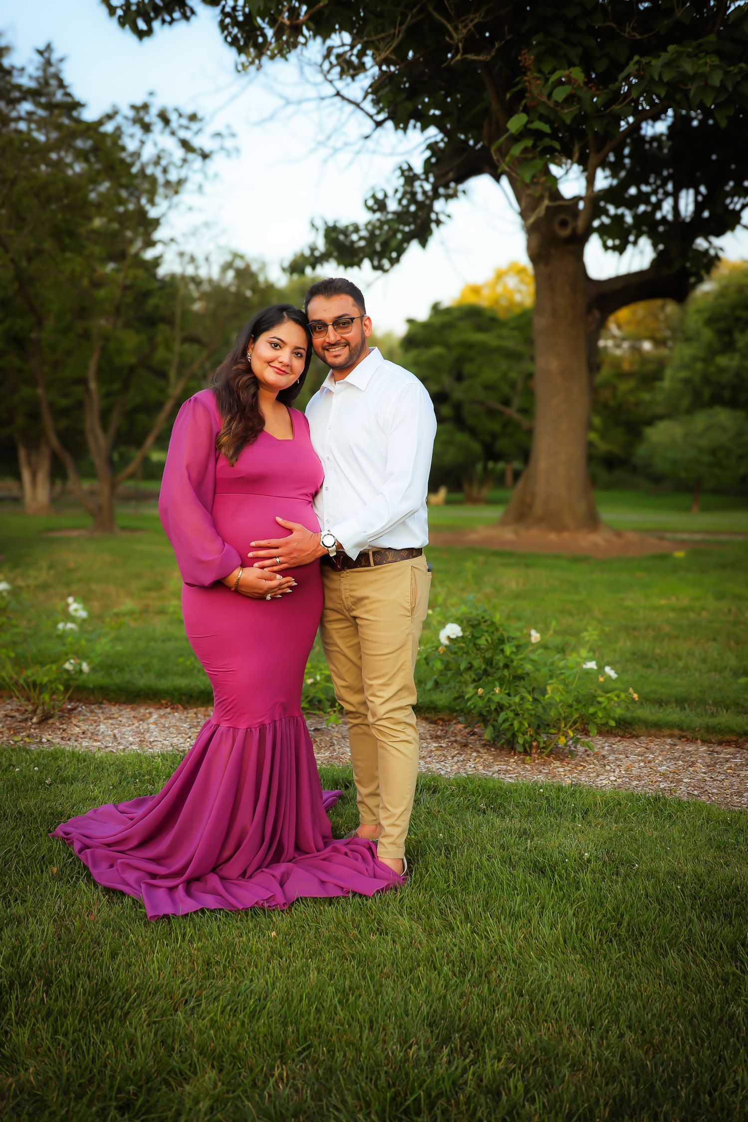 A couple in a park poses for maternity photos, with the woman wearing a flowing magenta dress and the man in khakis.