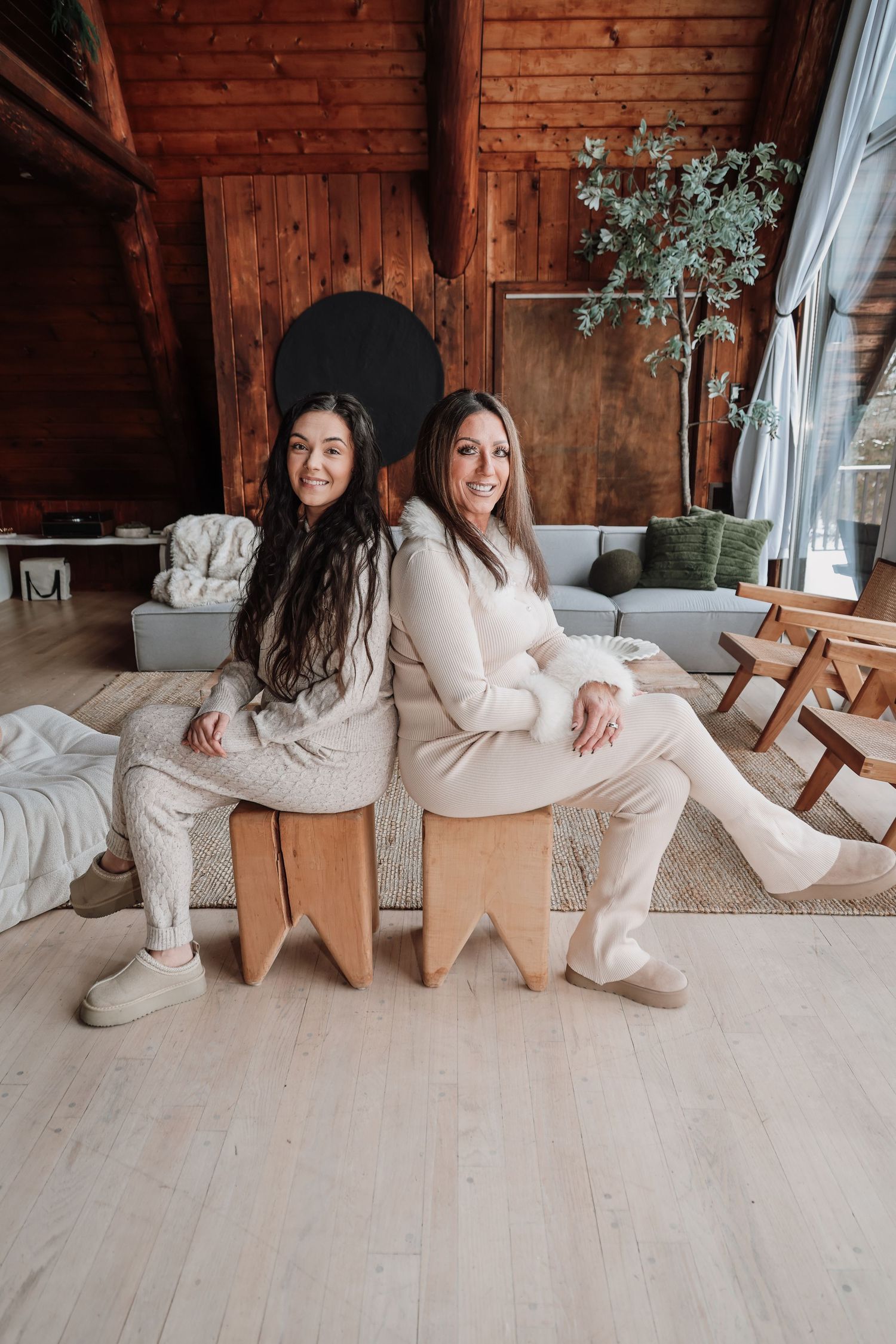 Two people in cozy beige loungewear sit back-to-back on wooden stools in a rustic cabin with wood paneled walls.
