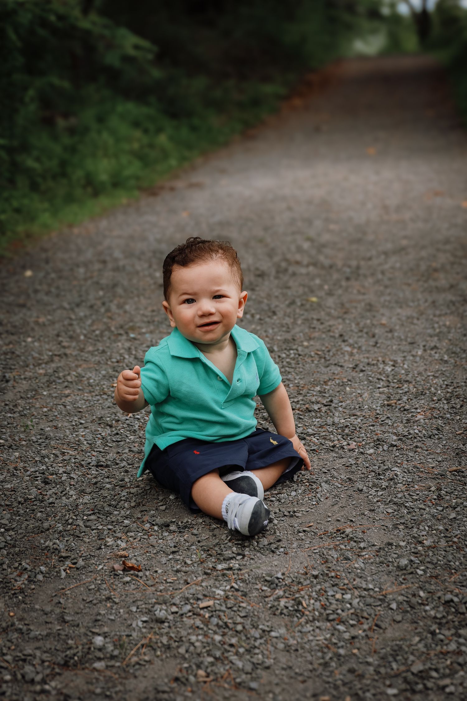 Small child in teal polo shirt and navy shorts sits on a gravel path giving thumbs up.