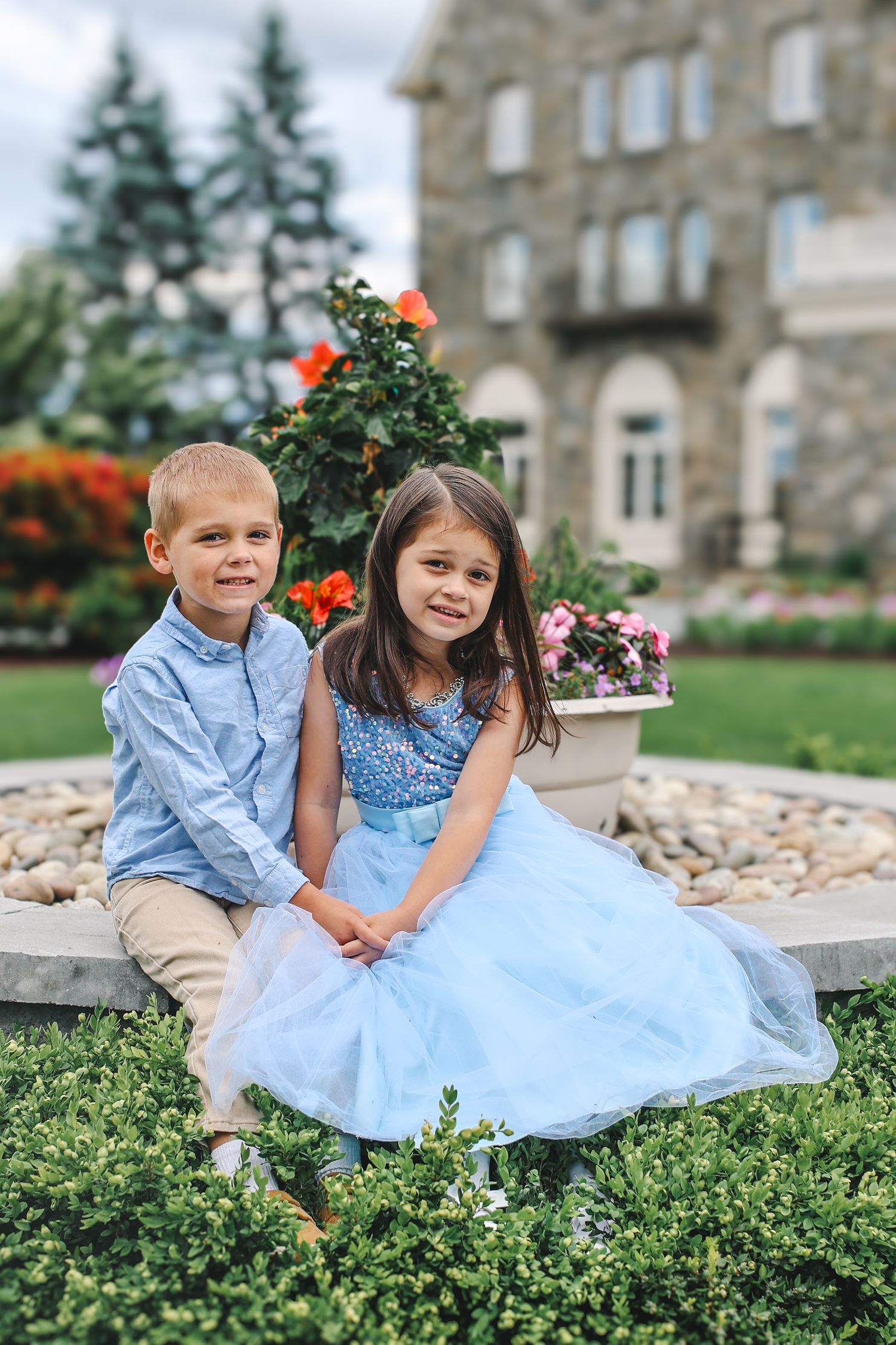 Two children sit together on stone steps surrounded by greenery and flowers in front of a historic building.